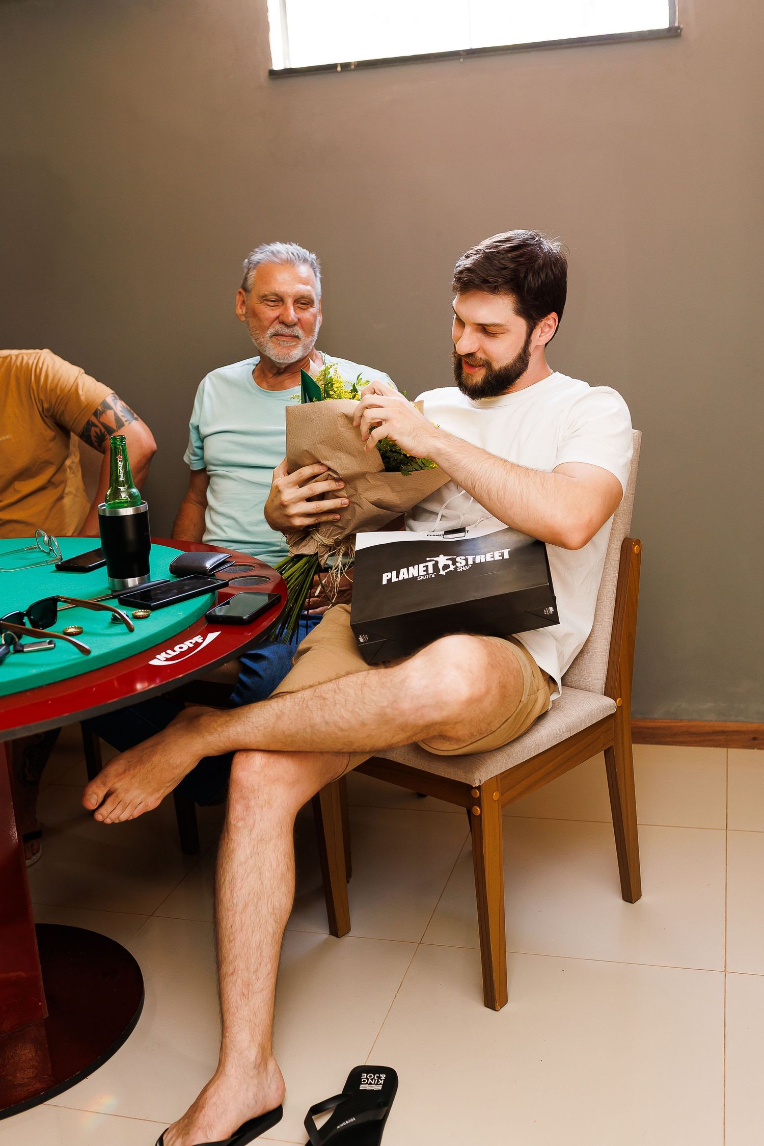 Two men sit at a green card table, one opening a Planet Fitness bag with groceries, while another smiles holding a beer bottle.