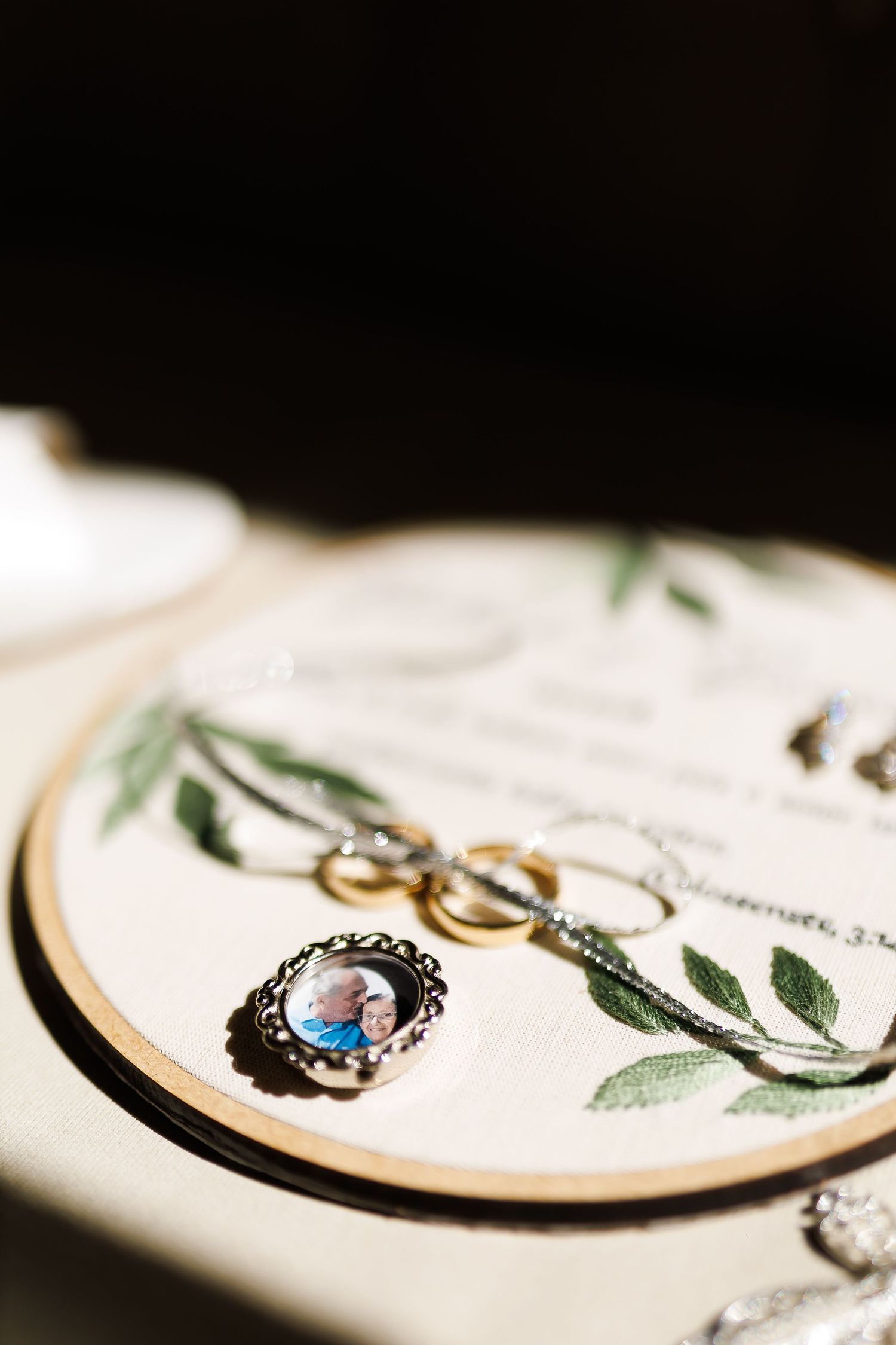 Wedding rings, necklace, and photo charm arranged on embroidered hoop with floral design for bridal flat lay.