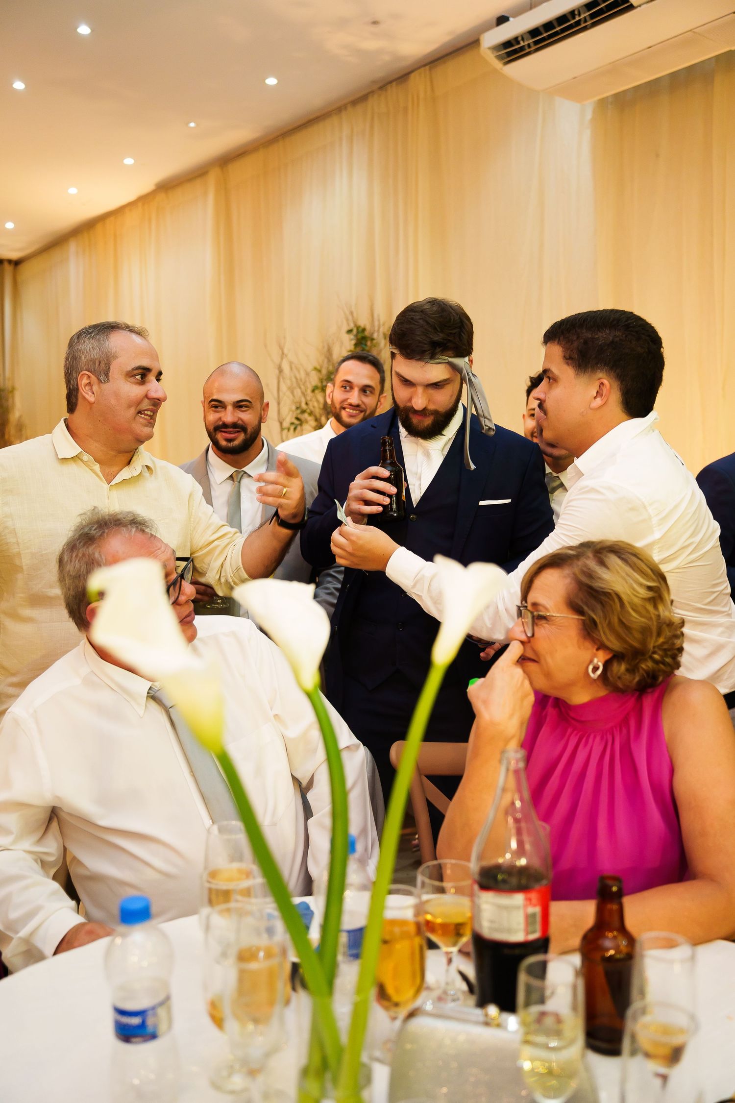 Groom surrounded by smiling guests at wedding reception dinner, with flowers and drinks on table.