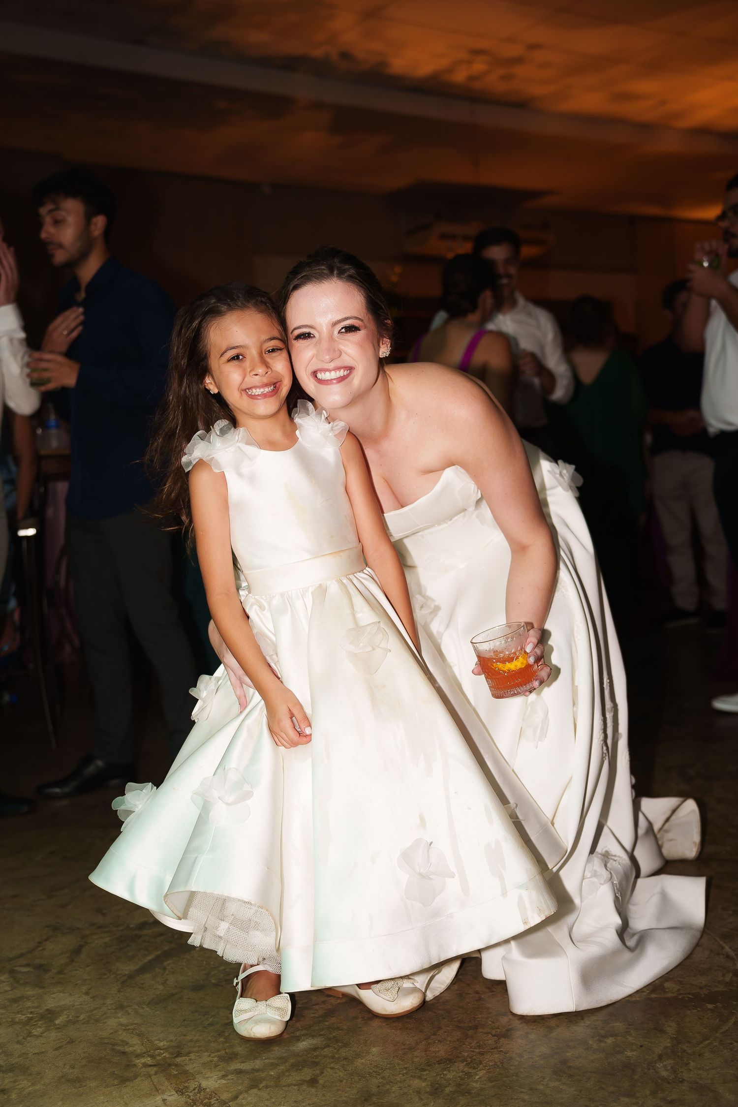 Bride in strapless white gown crouches beside flower girl in white dress, both smiling at wedding reception.