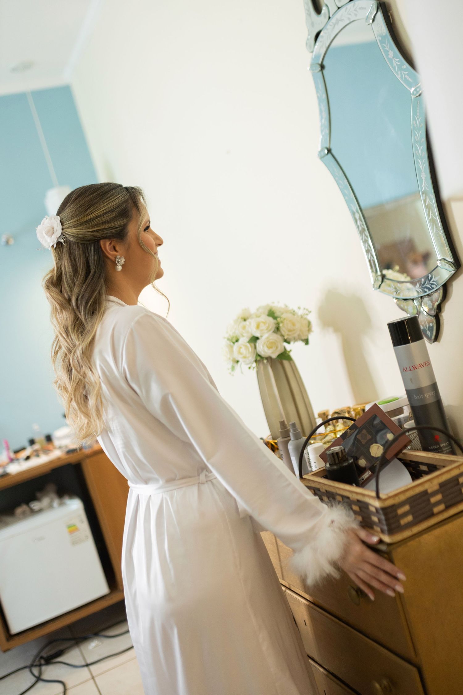 Bride in white robe with feather cuffs stands by wooden dresser with mirror and white flowers.
