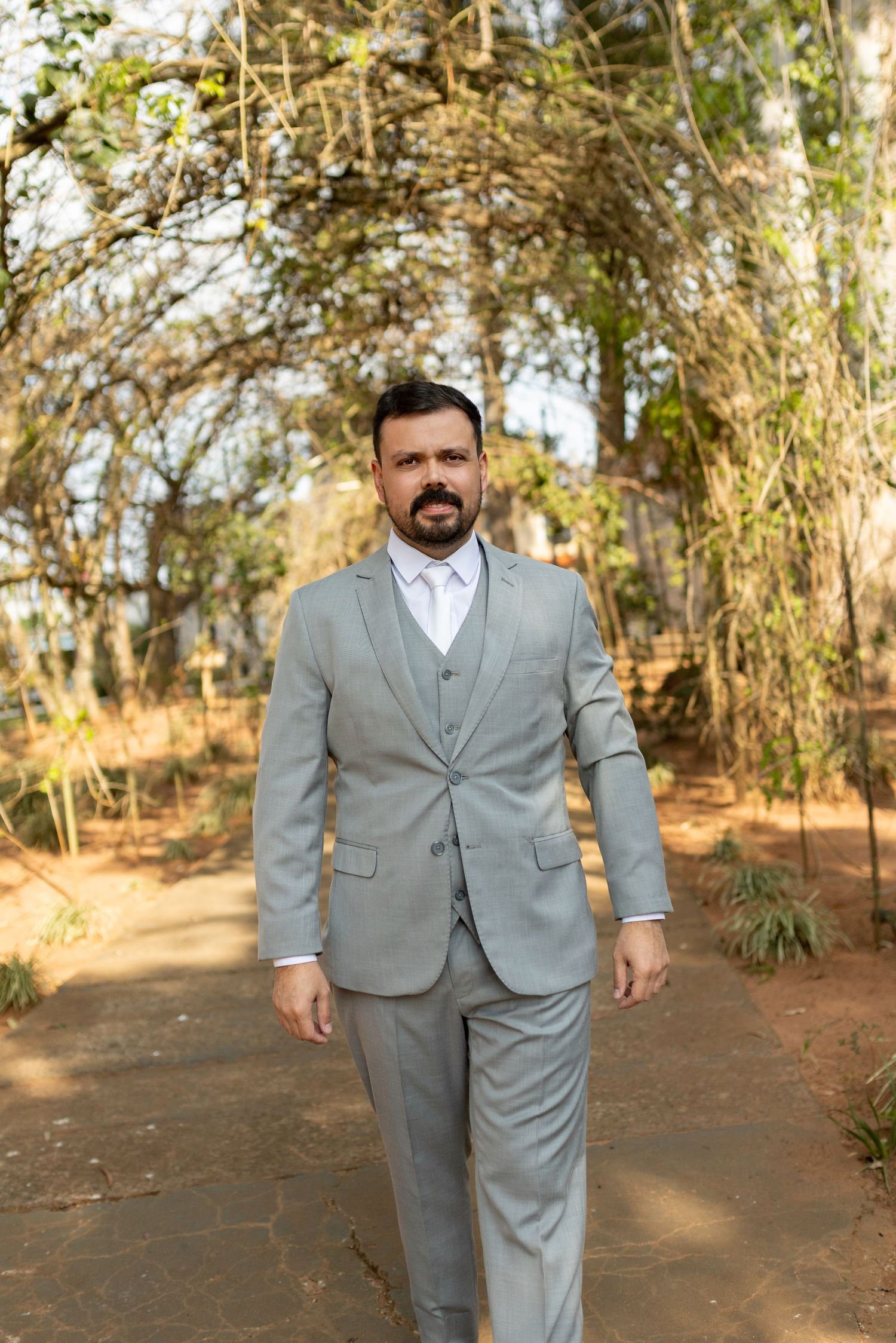 A well-dressed man in a light gray three-piece suit walks along a garden path surrounded by lush green trees.