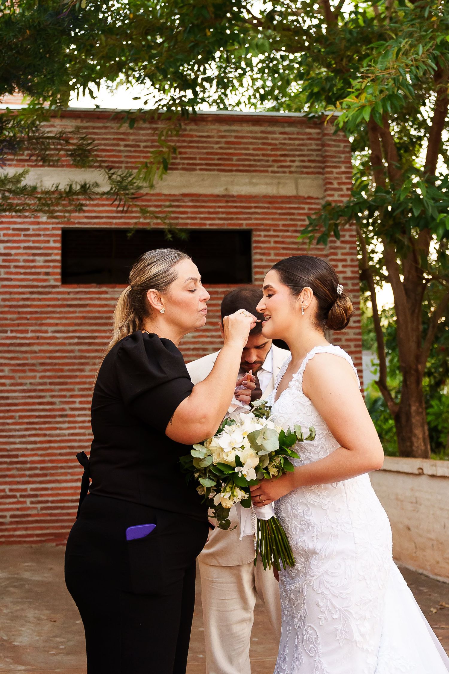 Makeup artist applying finishing touches to a bride in a white lace gown holding a white bouquet outdoors.