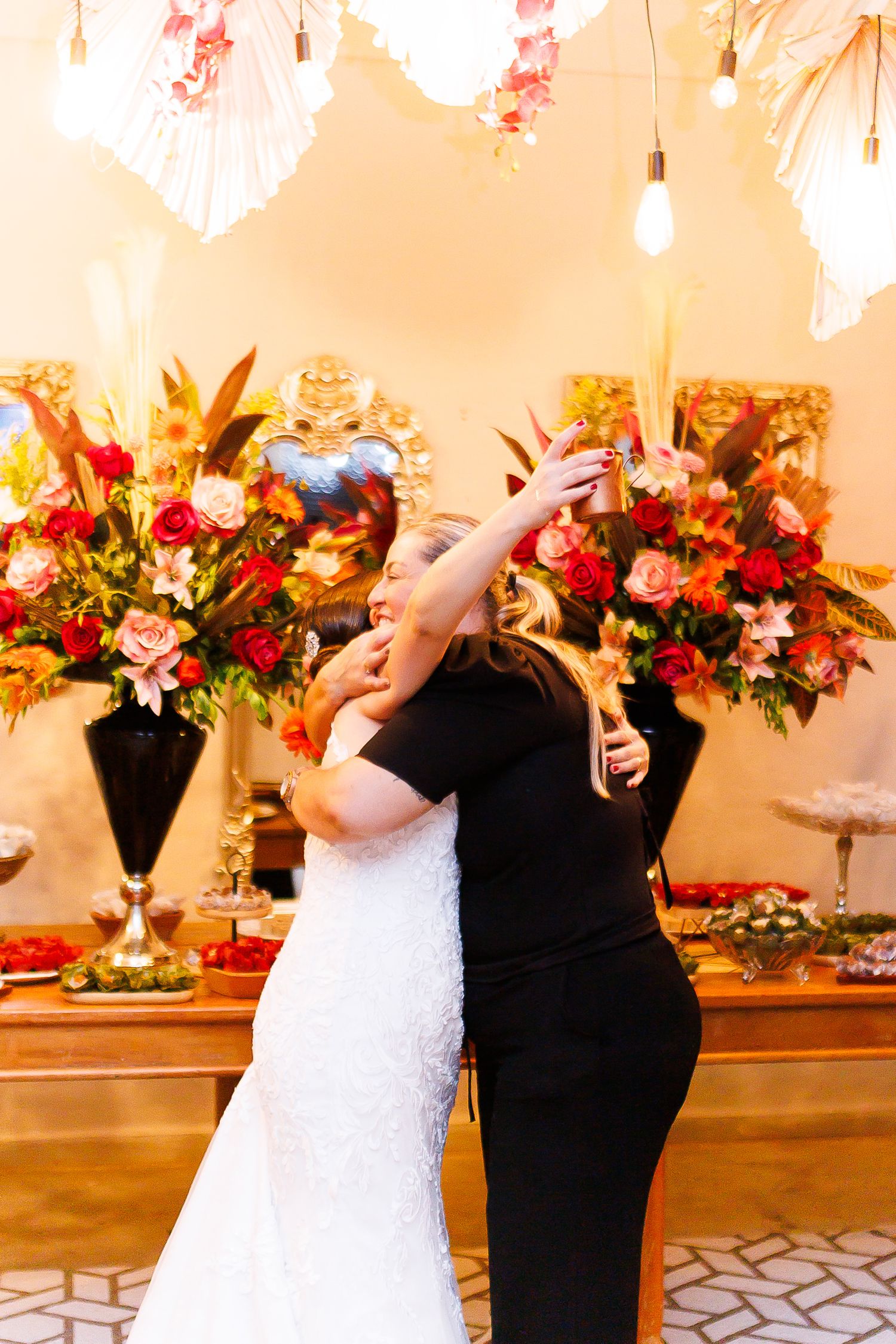 Bride in white lace gown embracing woman in black dress at elegant wedding reception with floral arrangements.