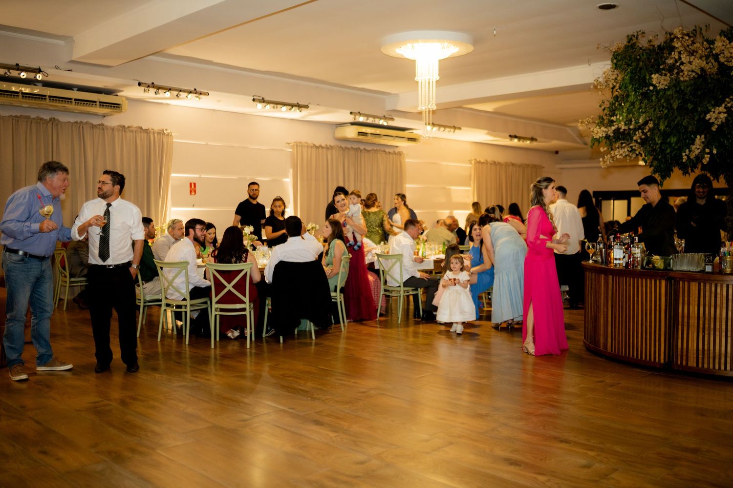 Elegant wedding reception with guests mingling on a wooden dance floor in a warmly lit banquet hall.