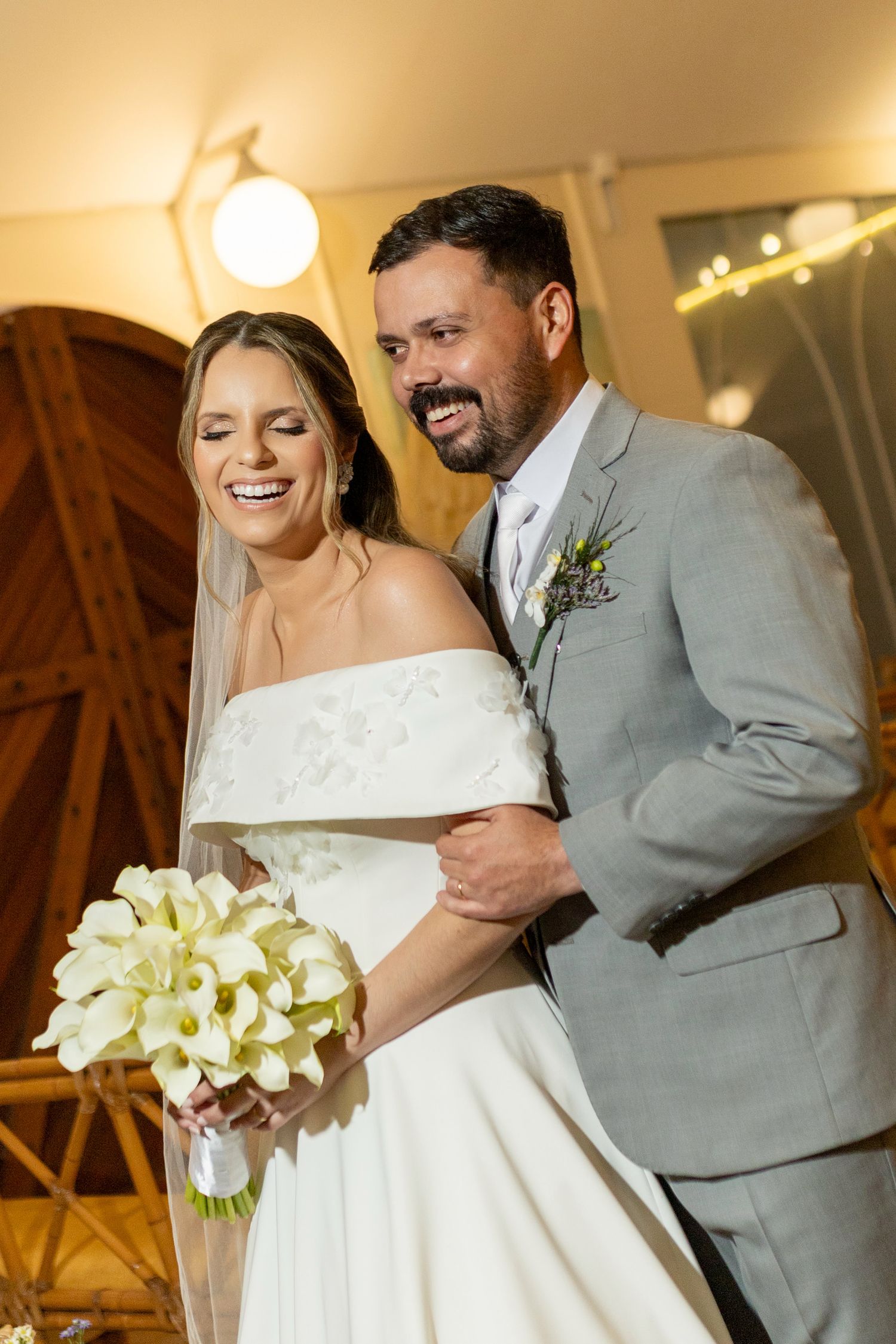 Happy bride in white off-shoulder gown holding calla lily bouquet stands with smiling groom in gray suit at wedding.
