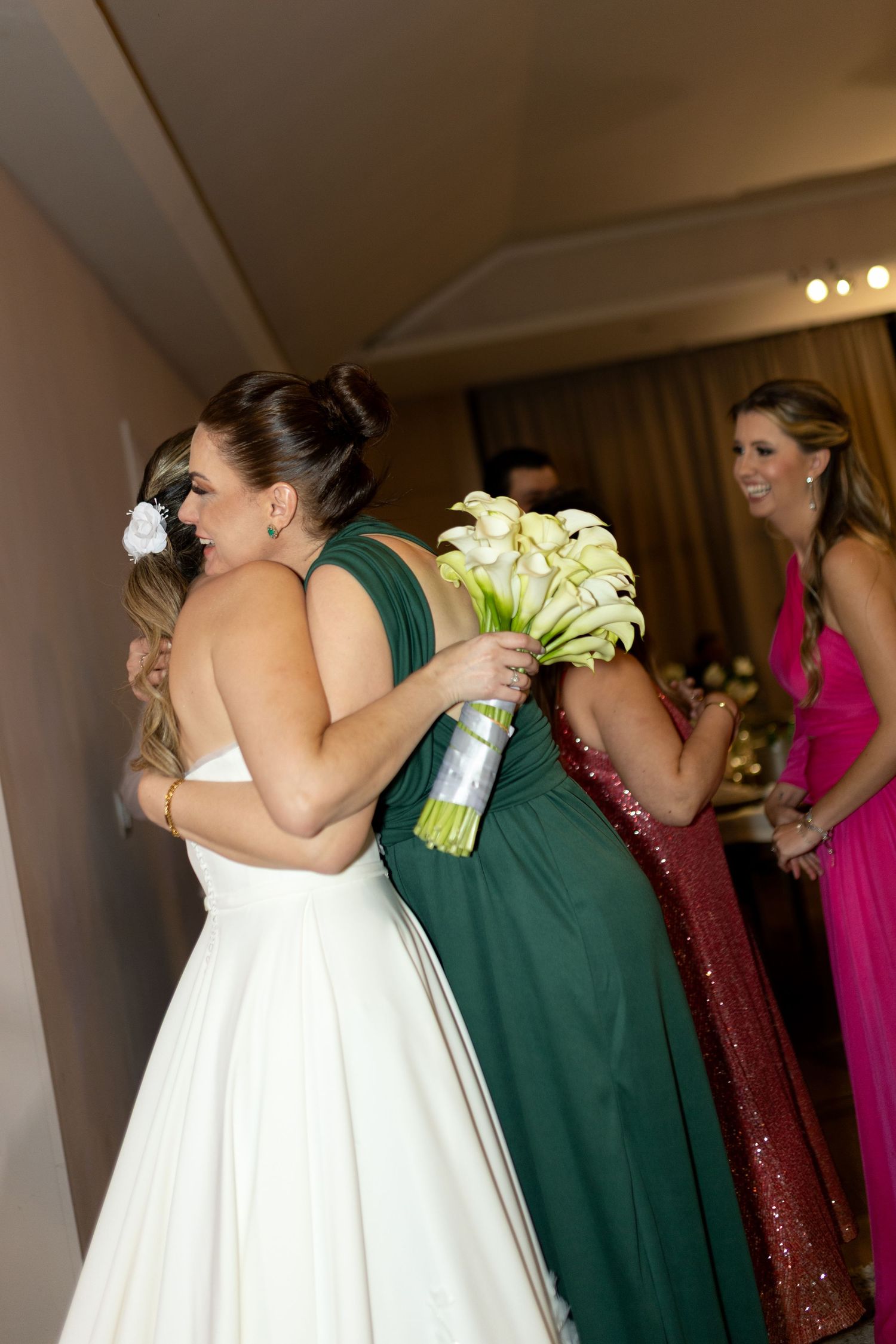 Bride in white gown hugging bridesmaid in green dress while holding white calla lily bouquet at wedding reception.