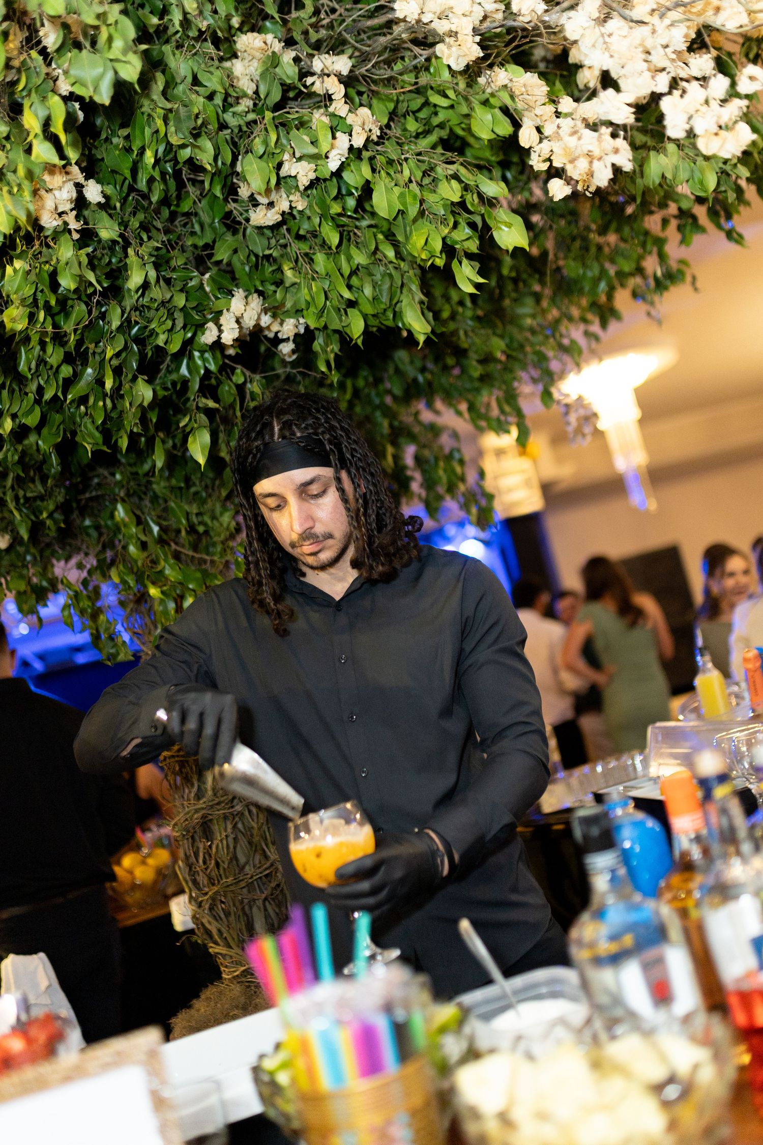 Bartender in black shirt pouring cocktail at decorated bar with lush green floral ceiling installation.