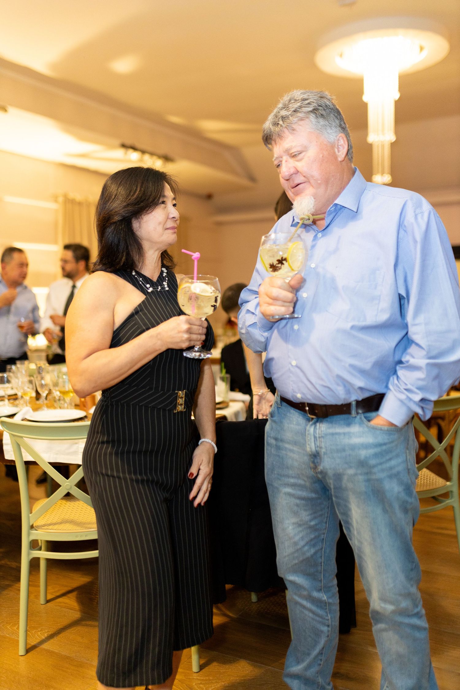 A man and woman in formal attire chat while holding cocktails at an elegant indoor social event.