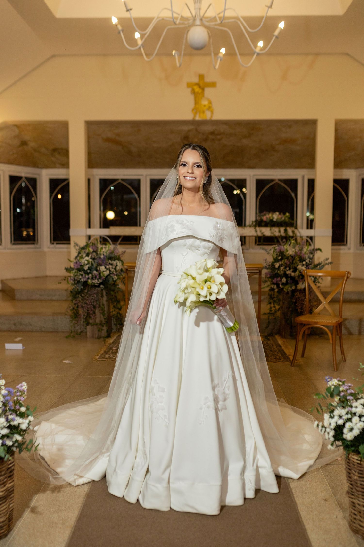 Bride in elegant off-shoulder white gown with veil holds bouquet inside decorated church with cross.