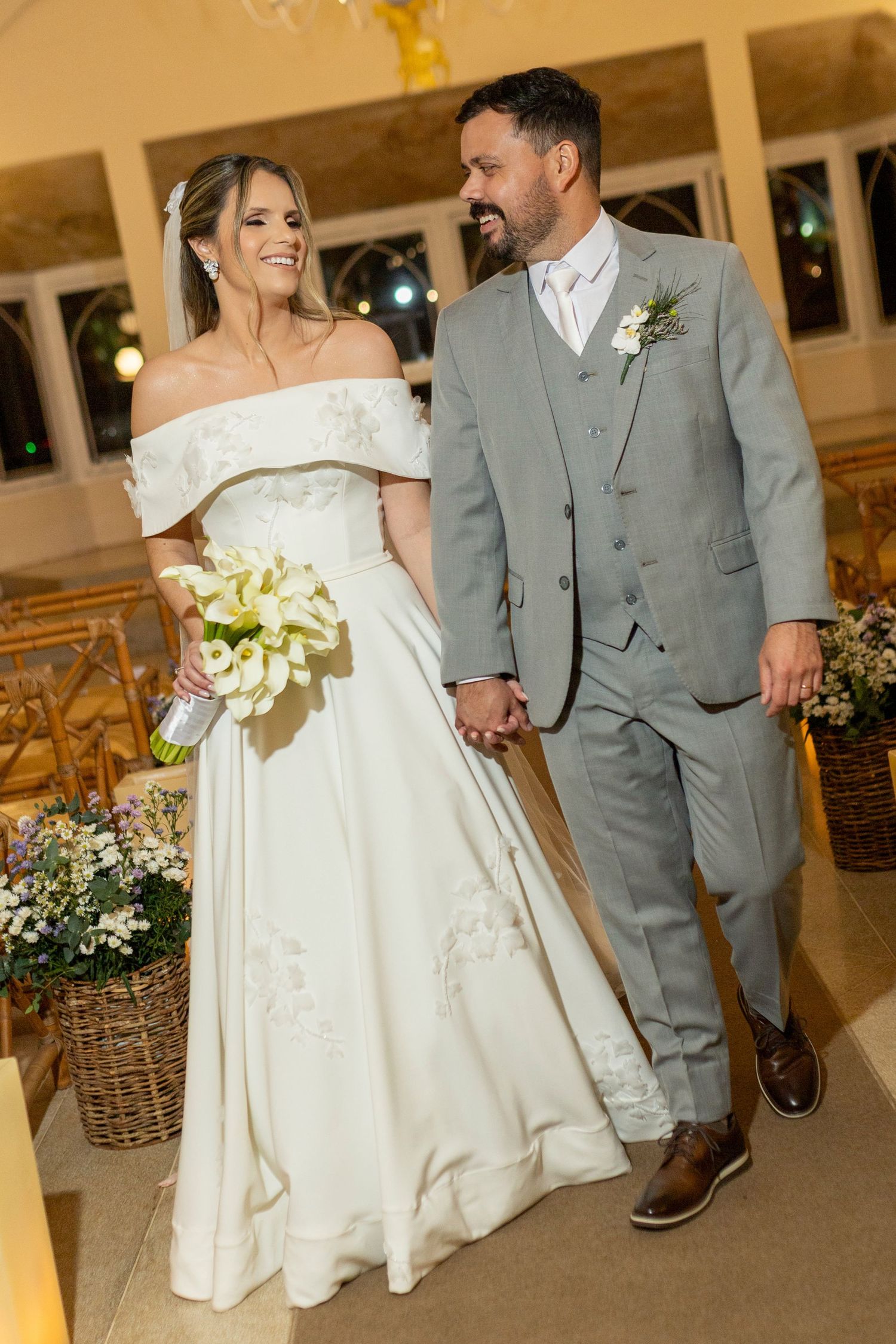 Bride in off-shoulder white gown holding bouquet walks with groom in gray suit down church aisle.
