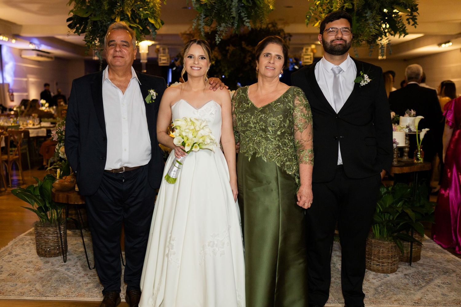 Bride in white gown poses with family at elegant wedding reception, surrounded by lush greenery decor.