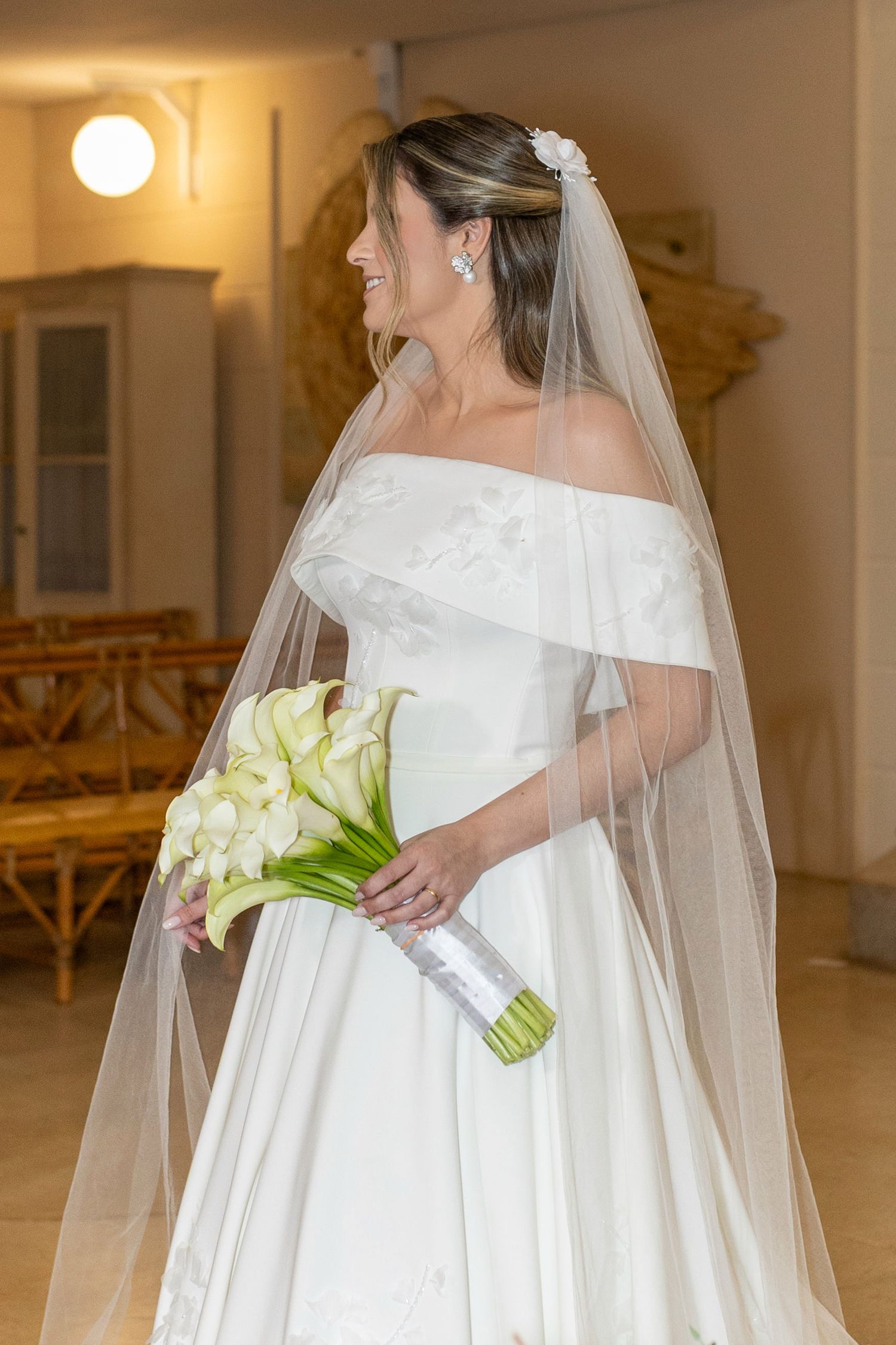 Elegant bride in white off-shoulder gown with veil holding calla lily bouquet, smiling indoors.