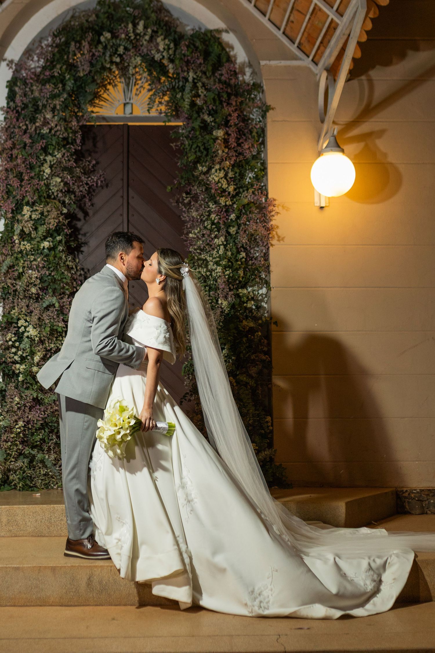 Bride and groom sharing a kiss on church steps at night, surrounded by floral arch and ivy-covered walls.