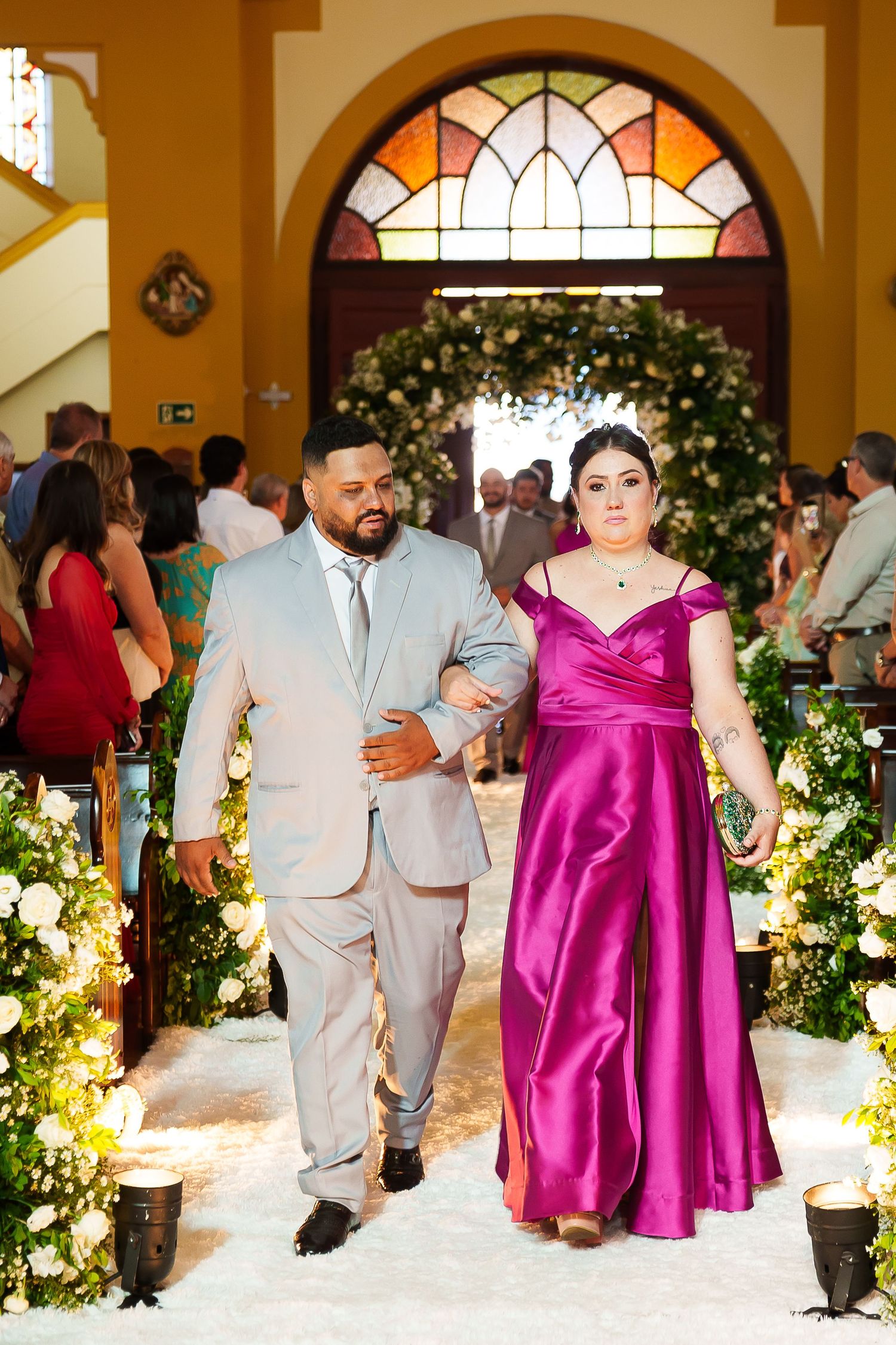 Couple walking down church aisle, man in gray suit, woman in magenta satin gown, floral arrangements, stained glass backdrop.