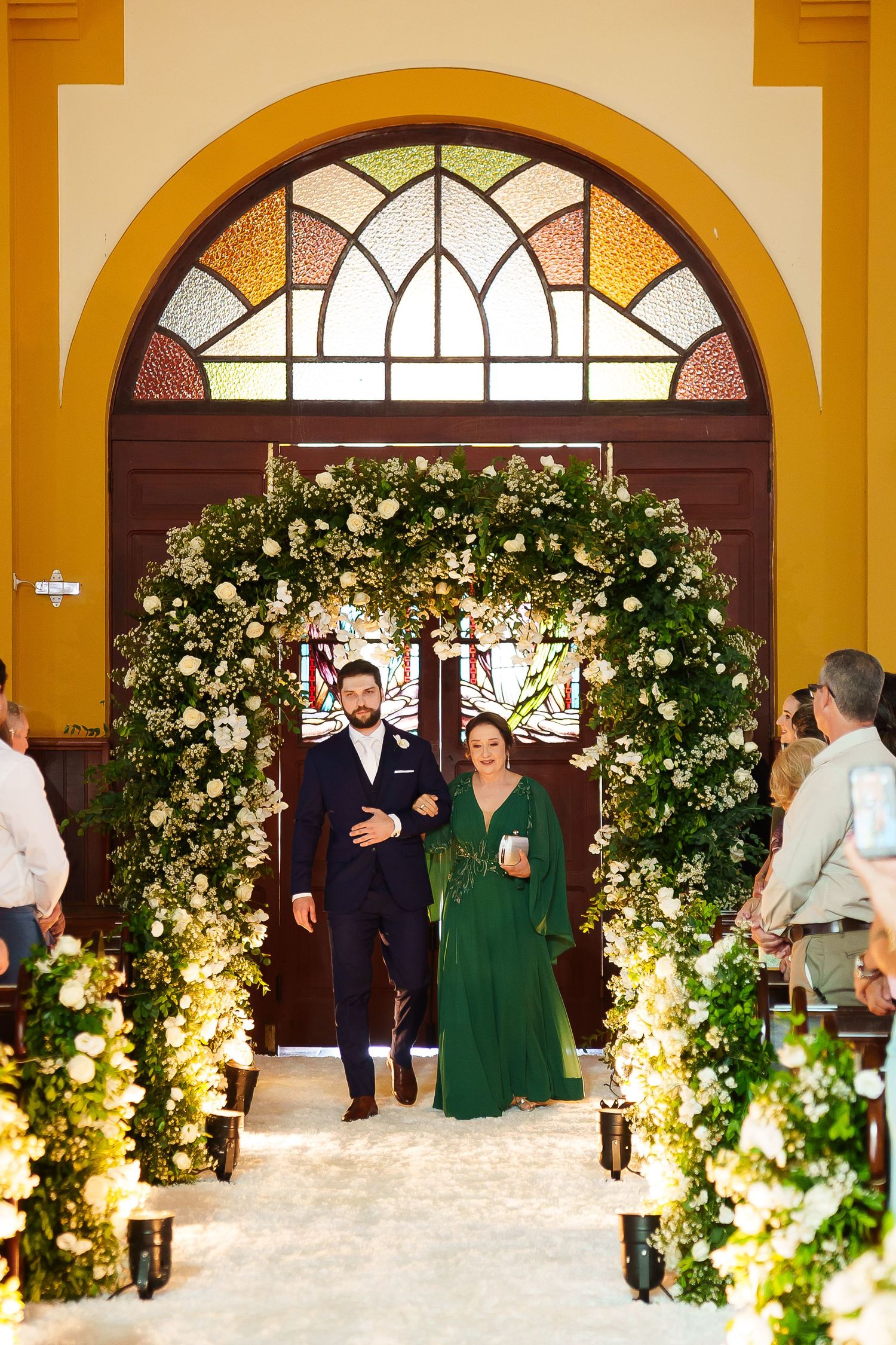 Couple walks through floral arch at church wedding ceremony, man in suit, woman in emerald green gown, candles lining aisle.