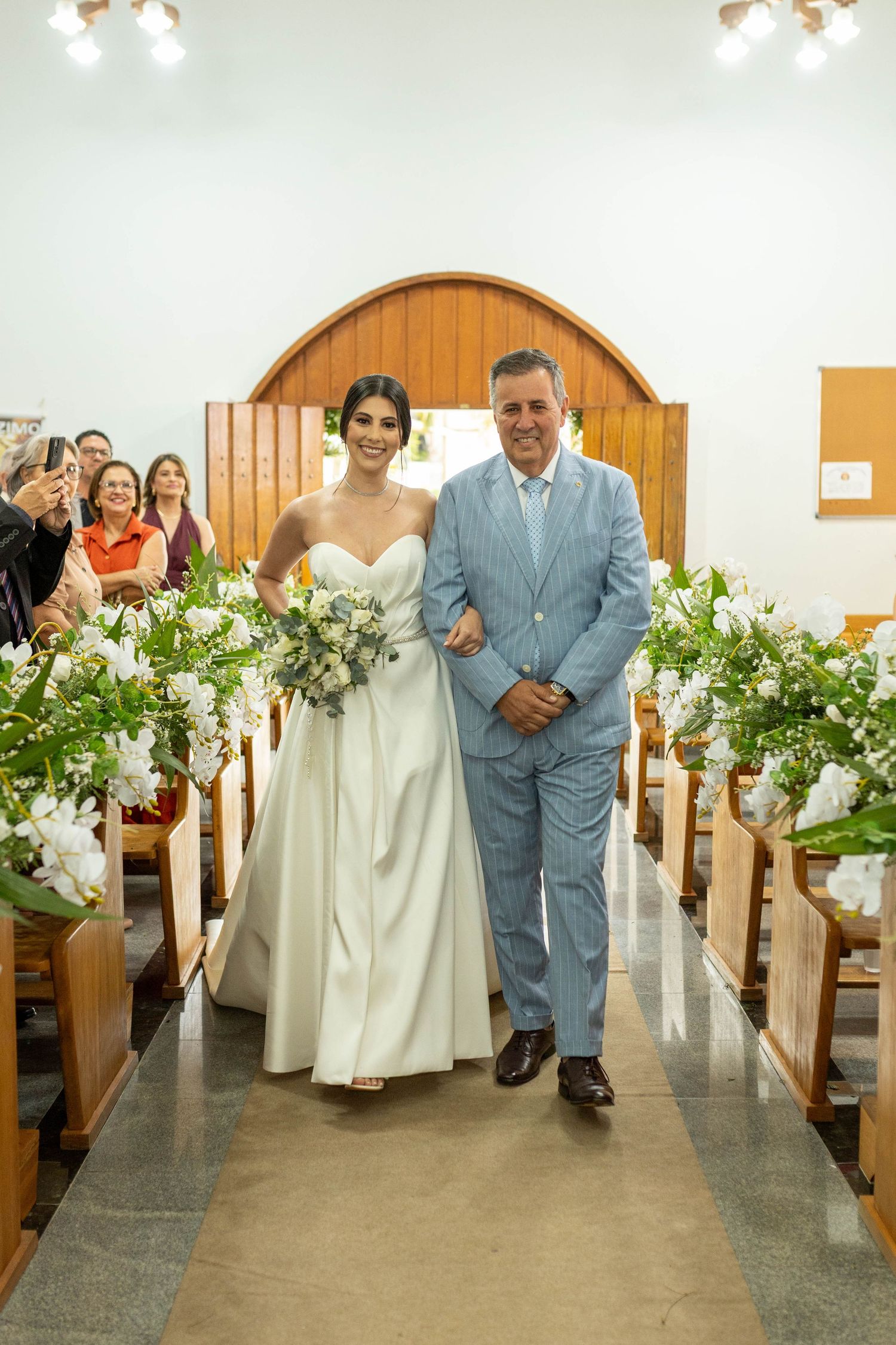 Bride in white gown walks down church aisle with escort, flanked by white floral arrangements on wooden pews.