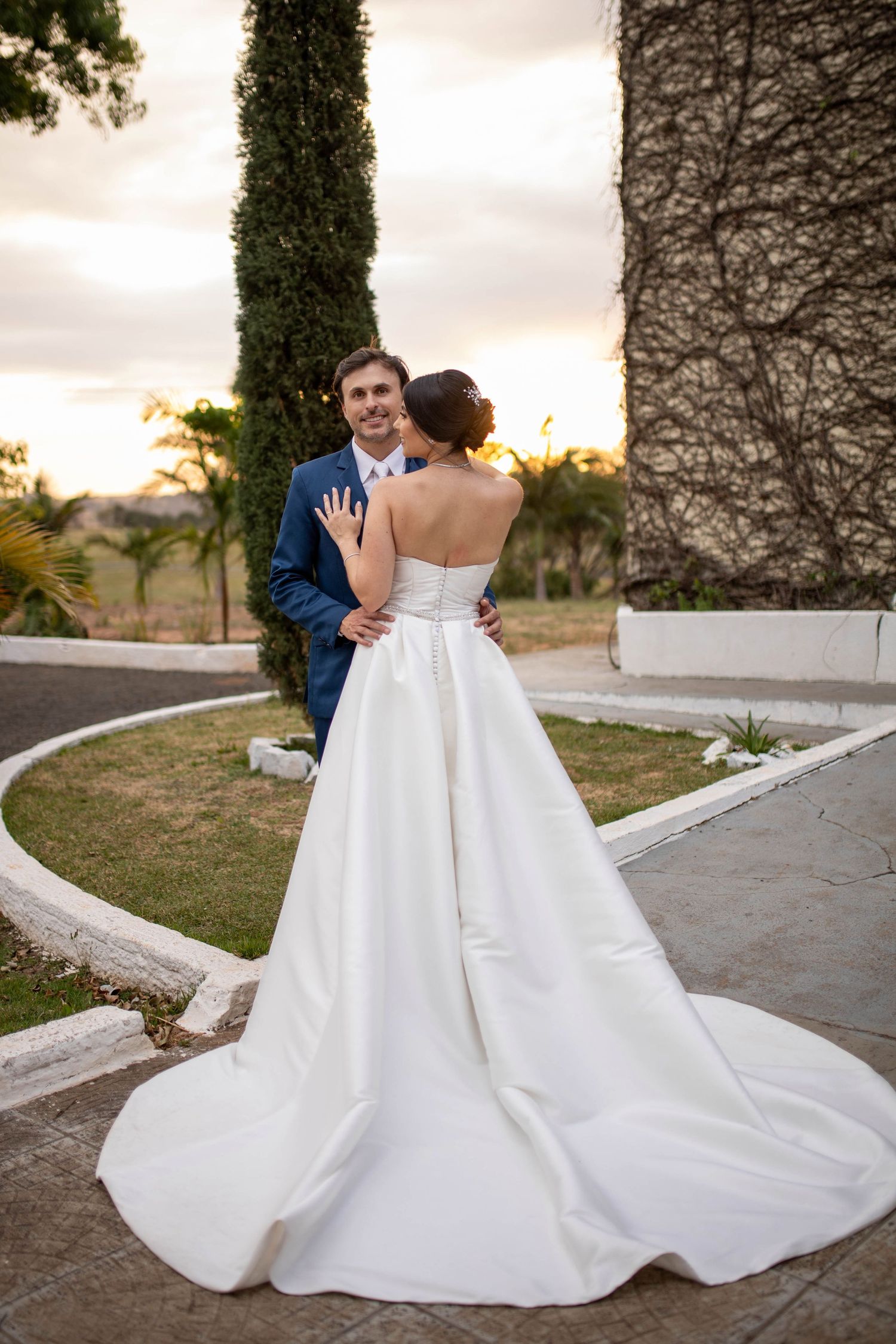 Bride in white strapless gown with train faces groom in blue suit outdoors at sunset near ivy-covered wall.
