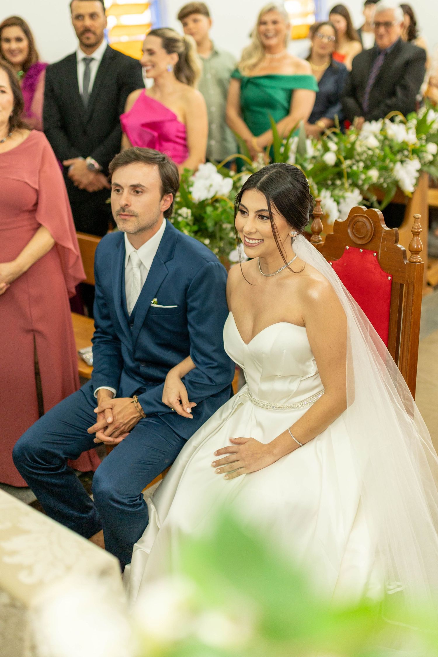 Bride in white gown and groom in blue suit sit together at church altar during wedding ceremony with guests in background.