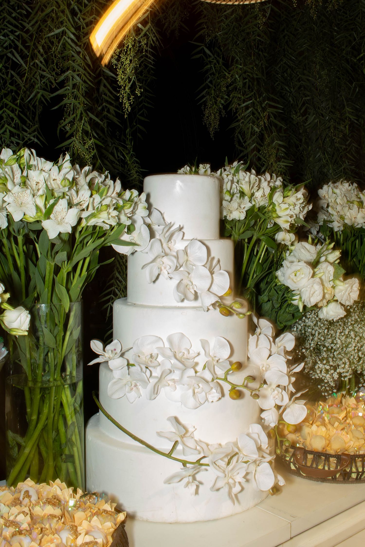 Elegant four-tier white wedding cake adorned with orchids, surrounded by white floral arrangements on a decorated table.