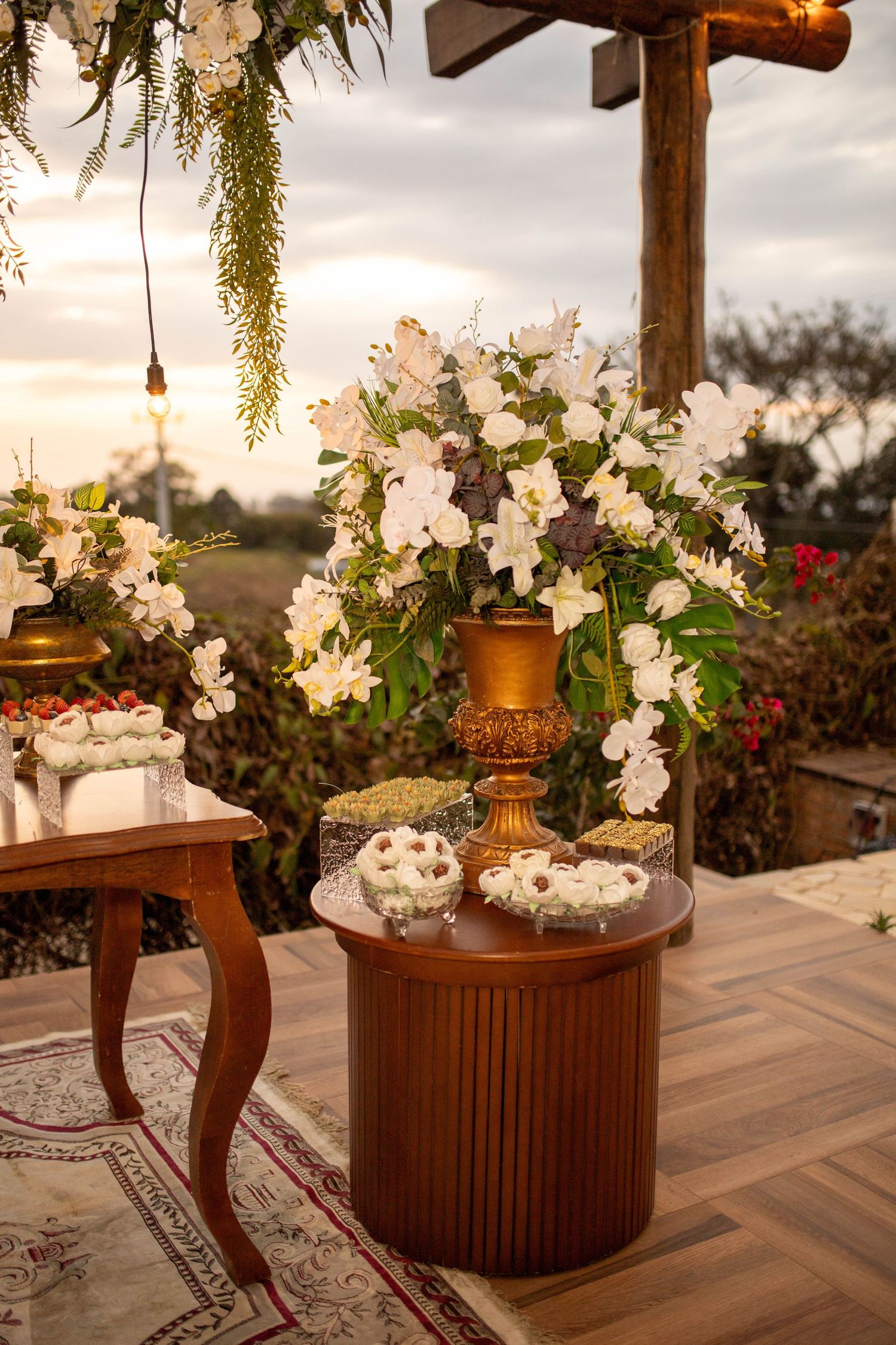 Elegant wedding dessert table with white floral arrangement in gold vase, wooden furniture, and sunset backdrop outdoors.