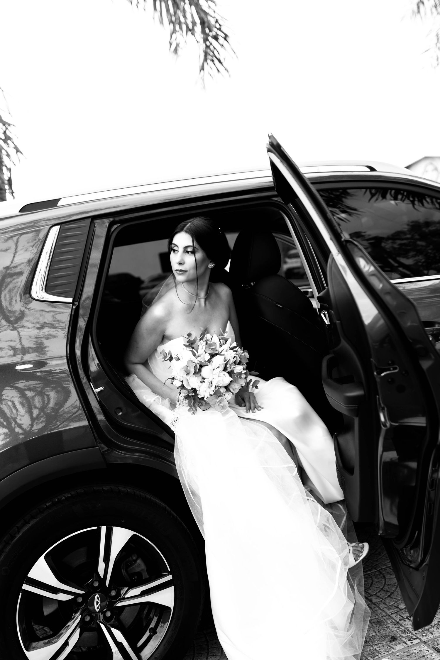 Black and white photo of a bride in a white gown holding a bouquet while sitting in a luxury car with door open.