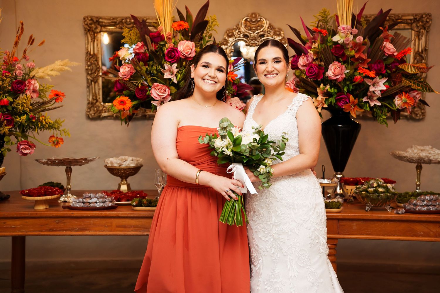 Bride in white lace gown and bridesmaid in coral dress pose together holding bouquet at elegant floral wedding reception.
