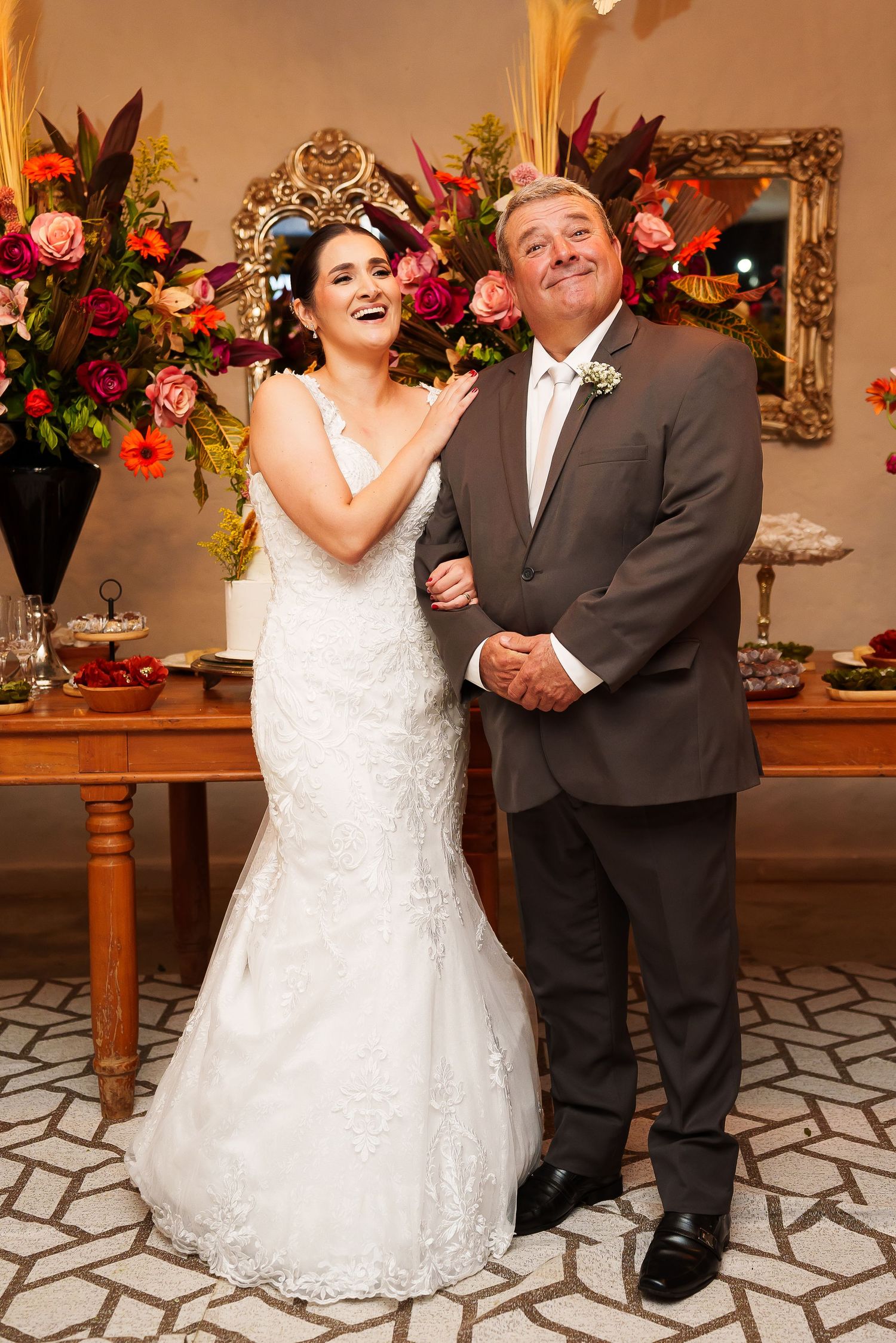 Bride in white lace gown laughing with older man in gray suit at elegant wedding reception with floral decor.