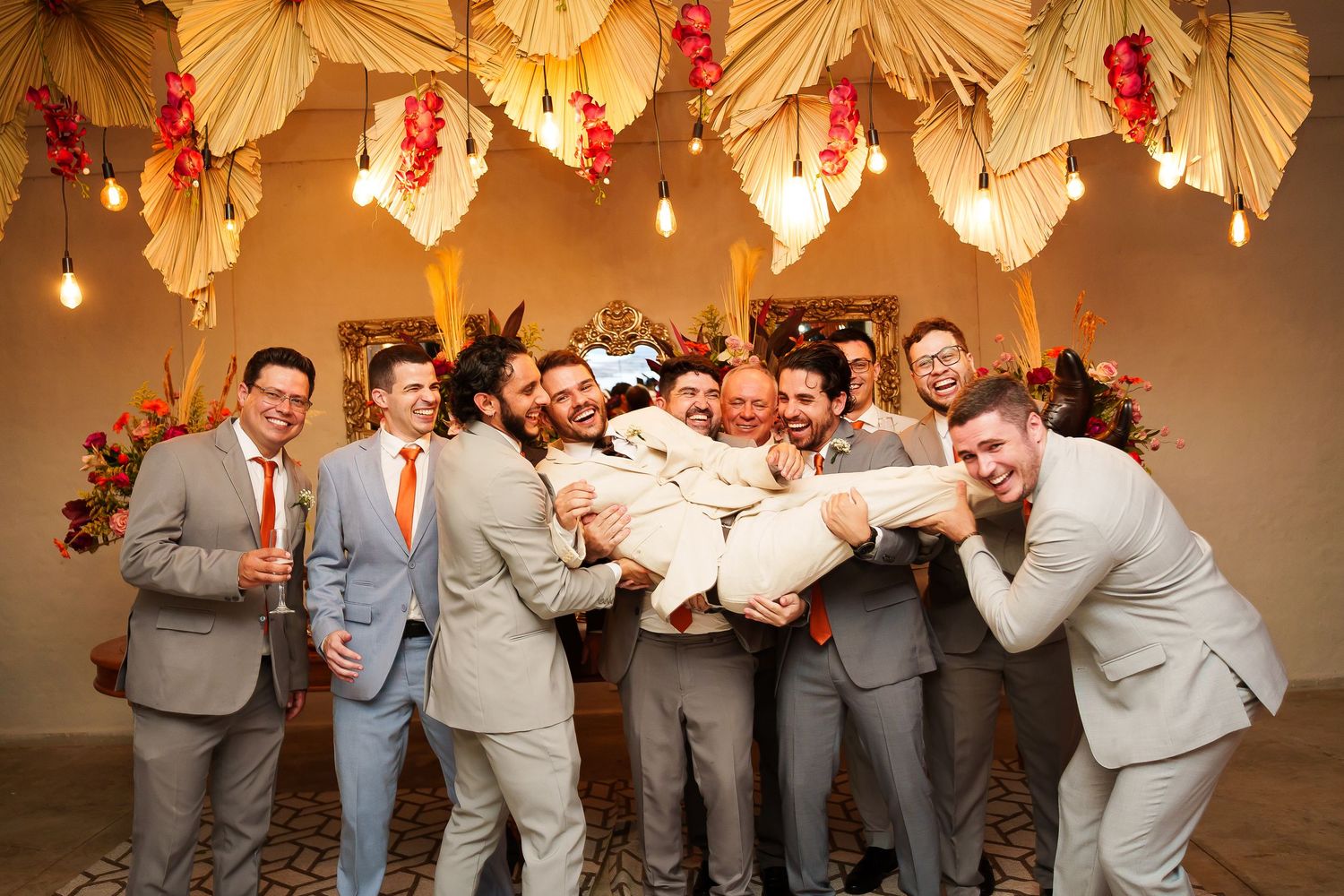 Groomsmen in tan suits lifting groom at wedding reception with floral ceiling decor and warm string lights.