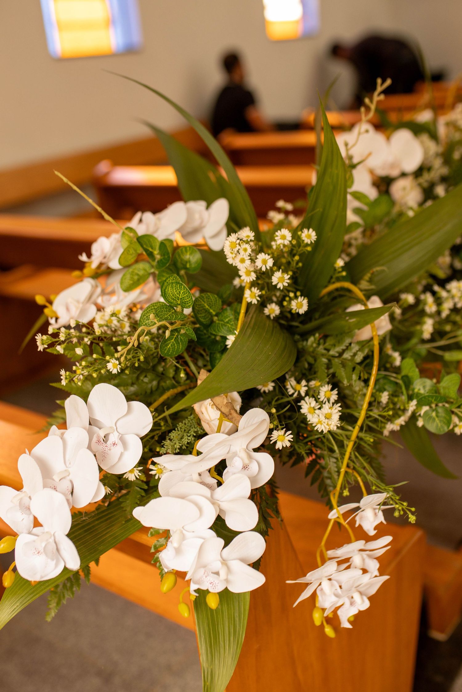 White orchid floral arrangement on wooden church pew with green foliage at a wedding ceremony.