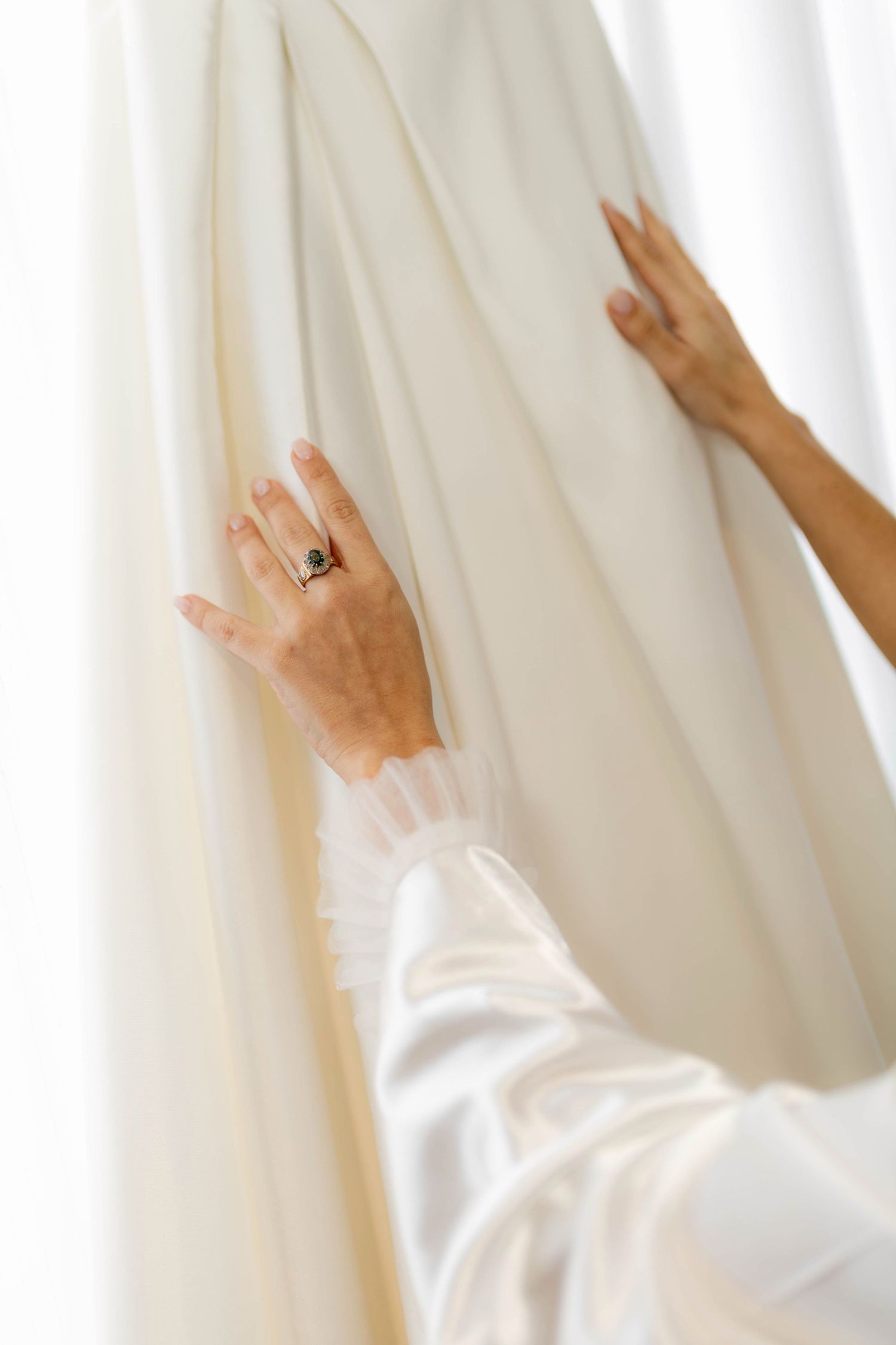 Bride touching ivory wedding dress fabric, wearing elegant ring on finger, soft white background.