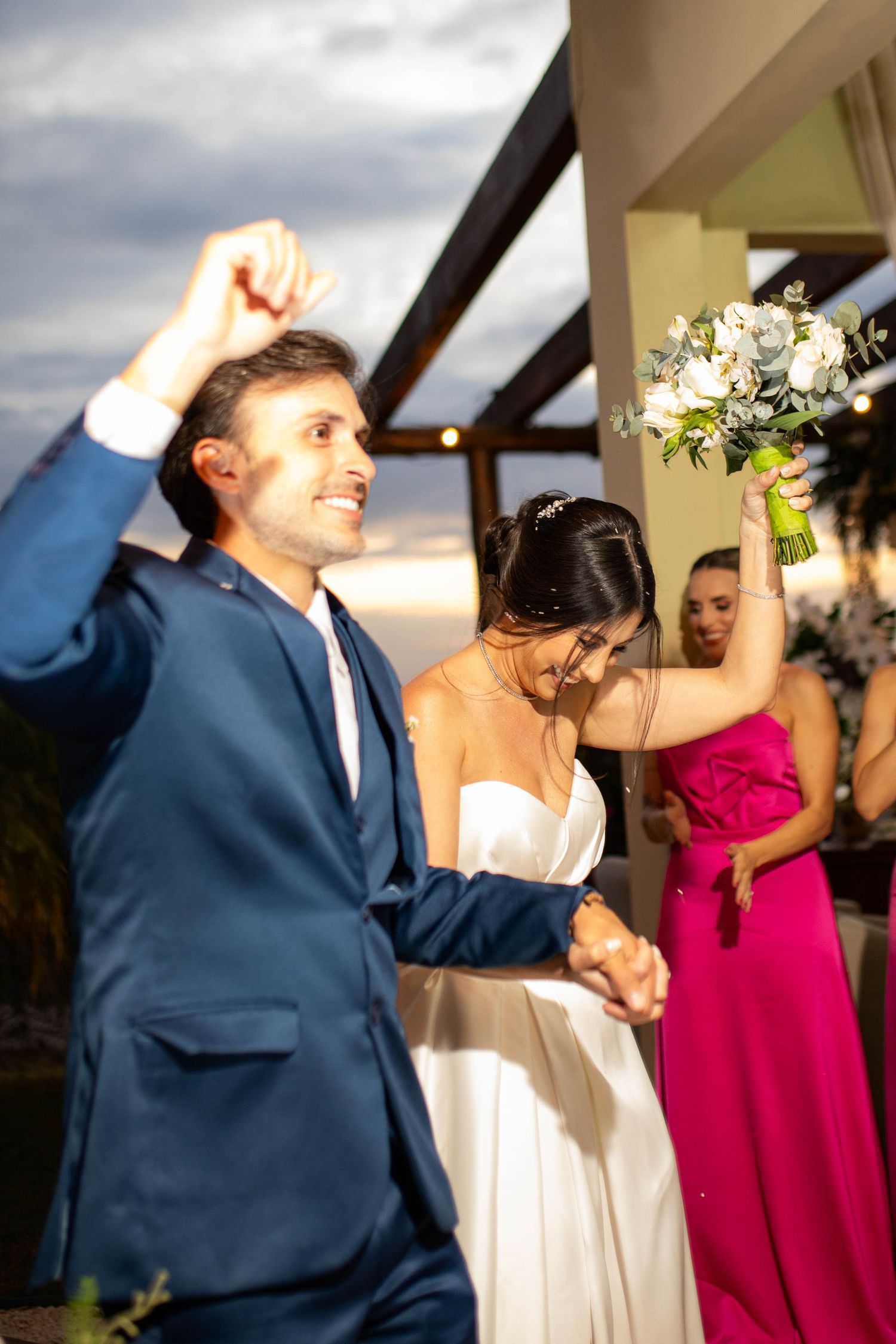 Joyful newlyweds celebrate at outdoor wedding reception, groom in blue suit raises fist as bride holds white bouquet.