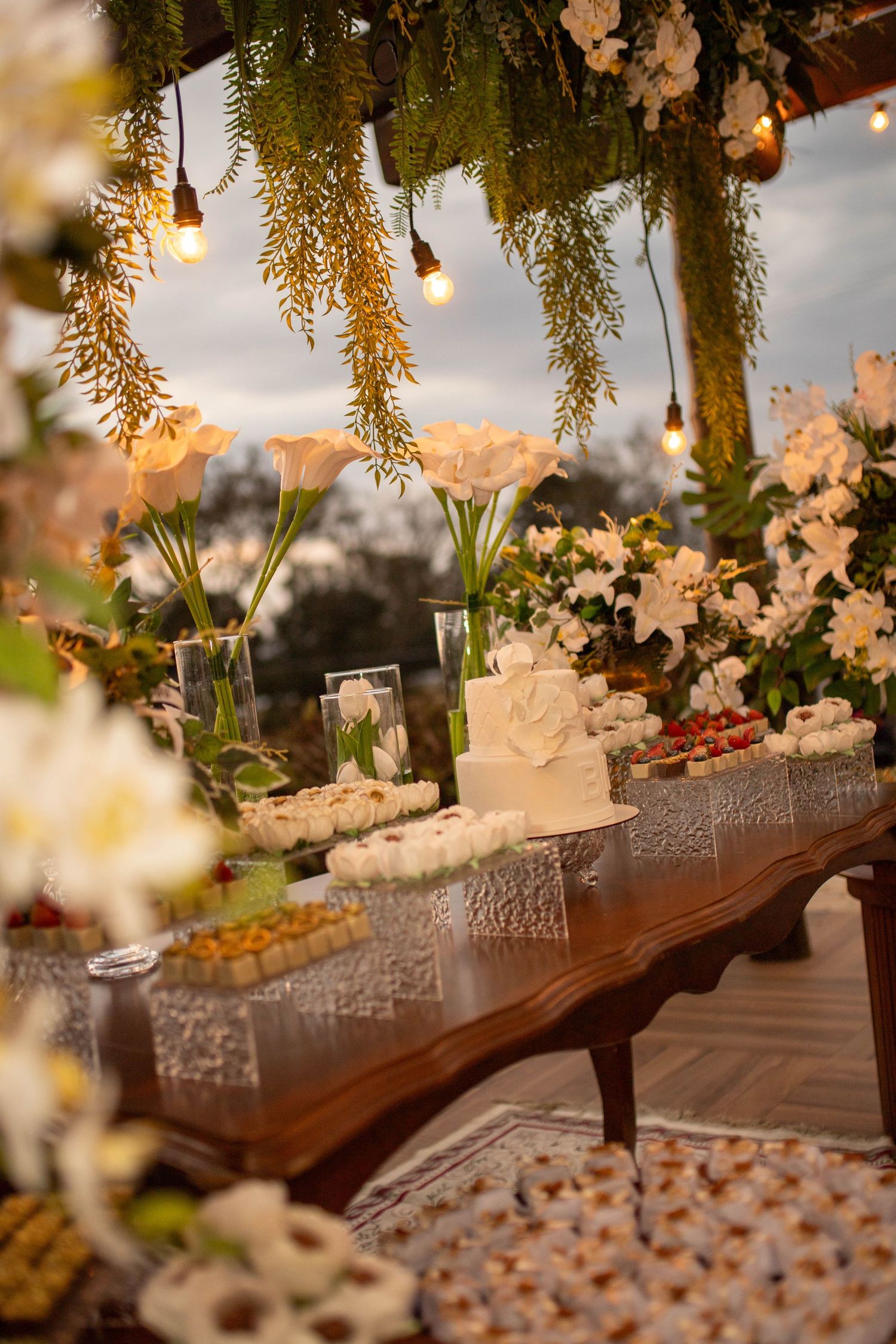 Elegant wedding dessert table with white cake, floral arrangements, and string lights at dusk.