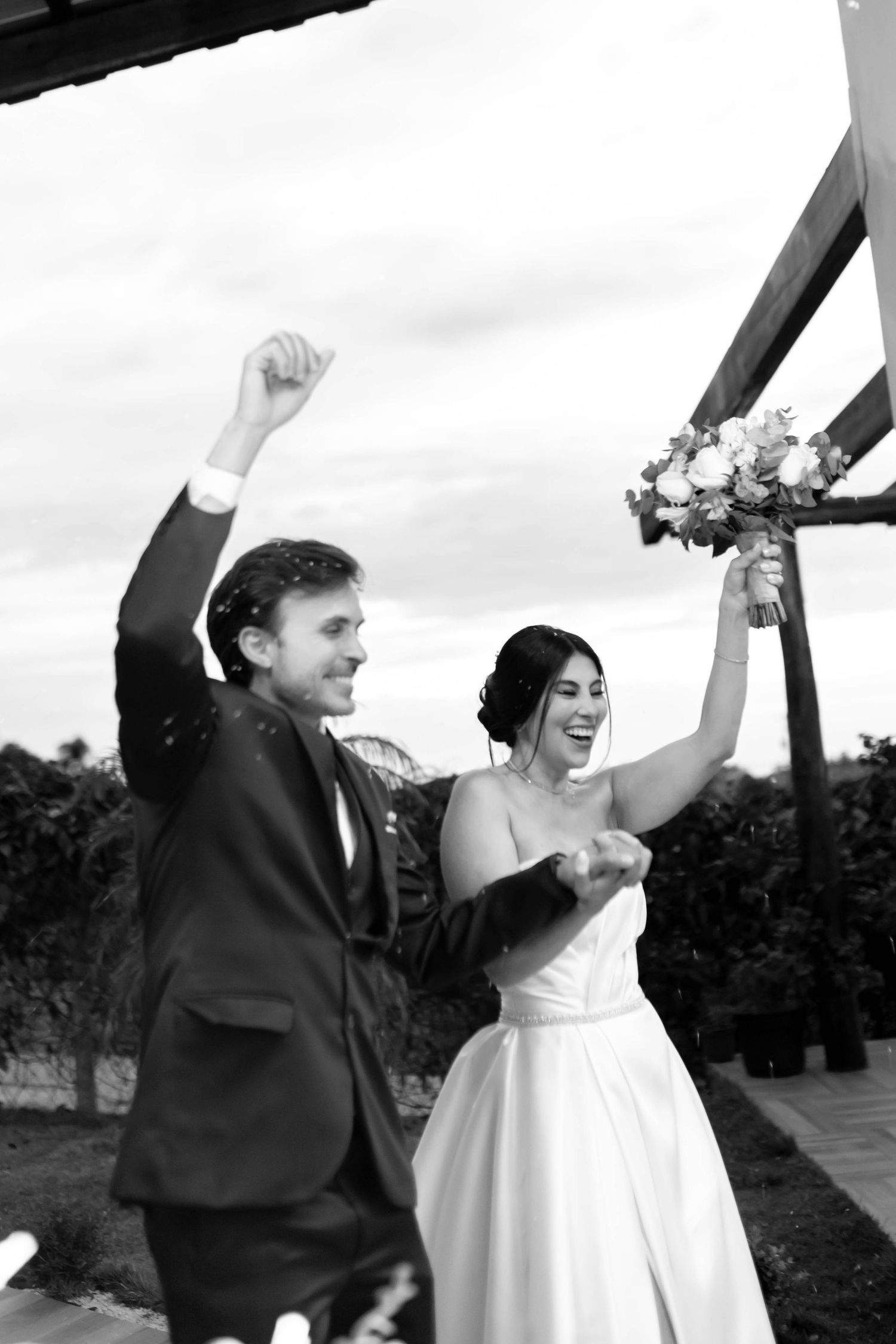 Black and white photo of joyful newlyweds celebrating after wedding ceremony, bride holding bouquet aloft.