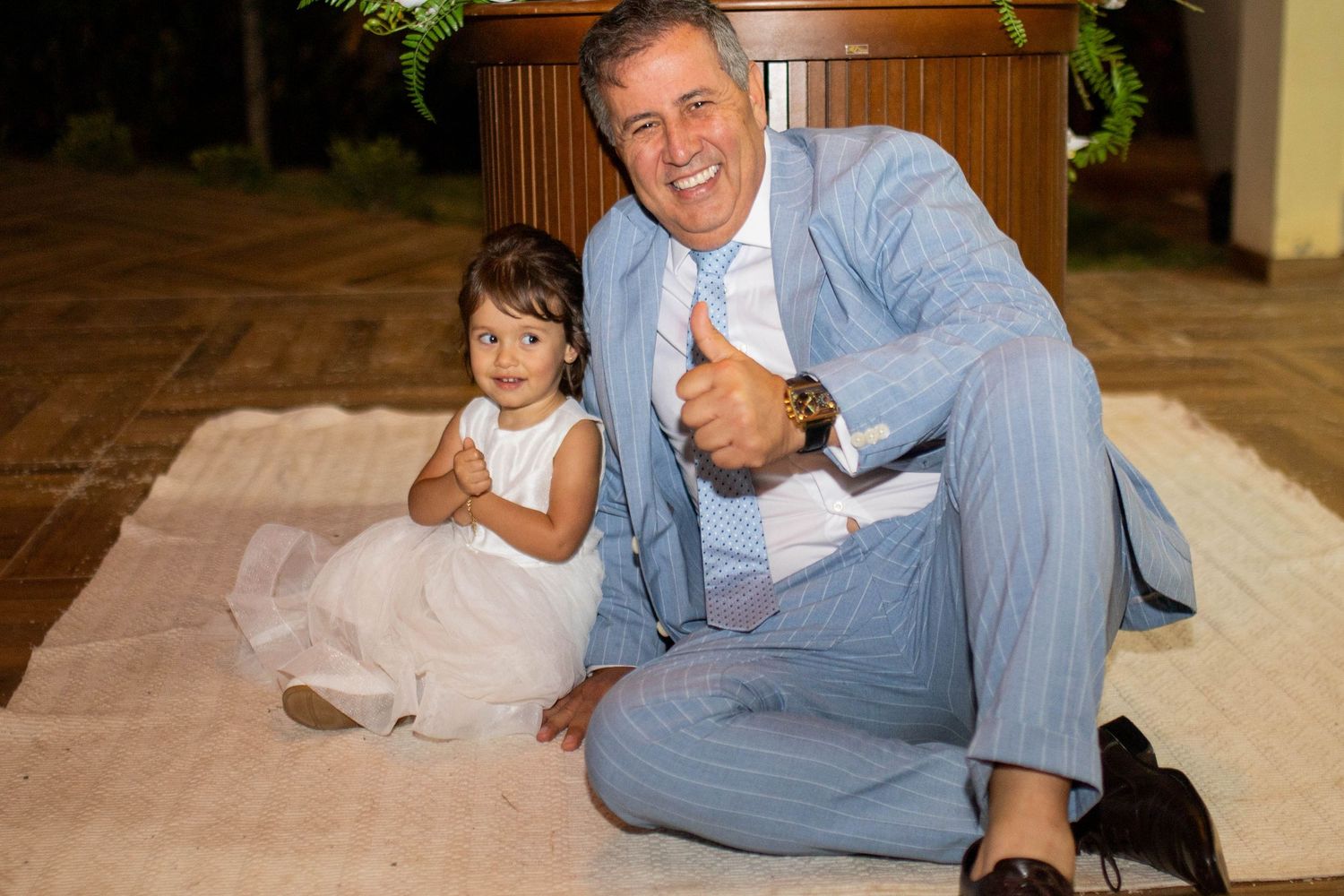 Man in blue suit gives thumbs up sitting on rug with young girl in white dress at elegant outdoor event.