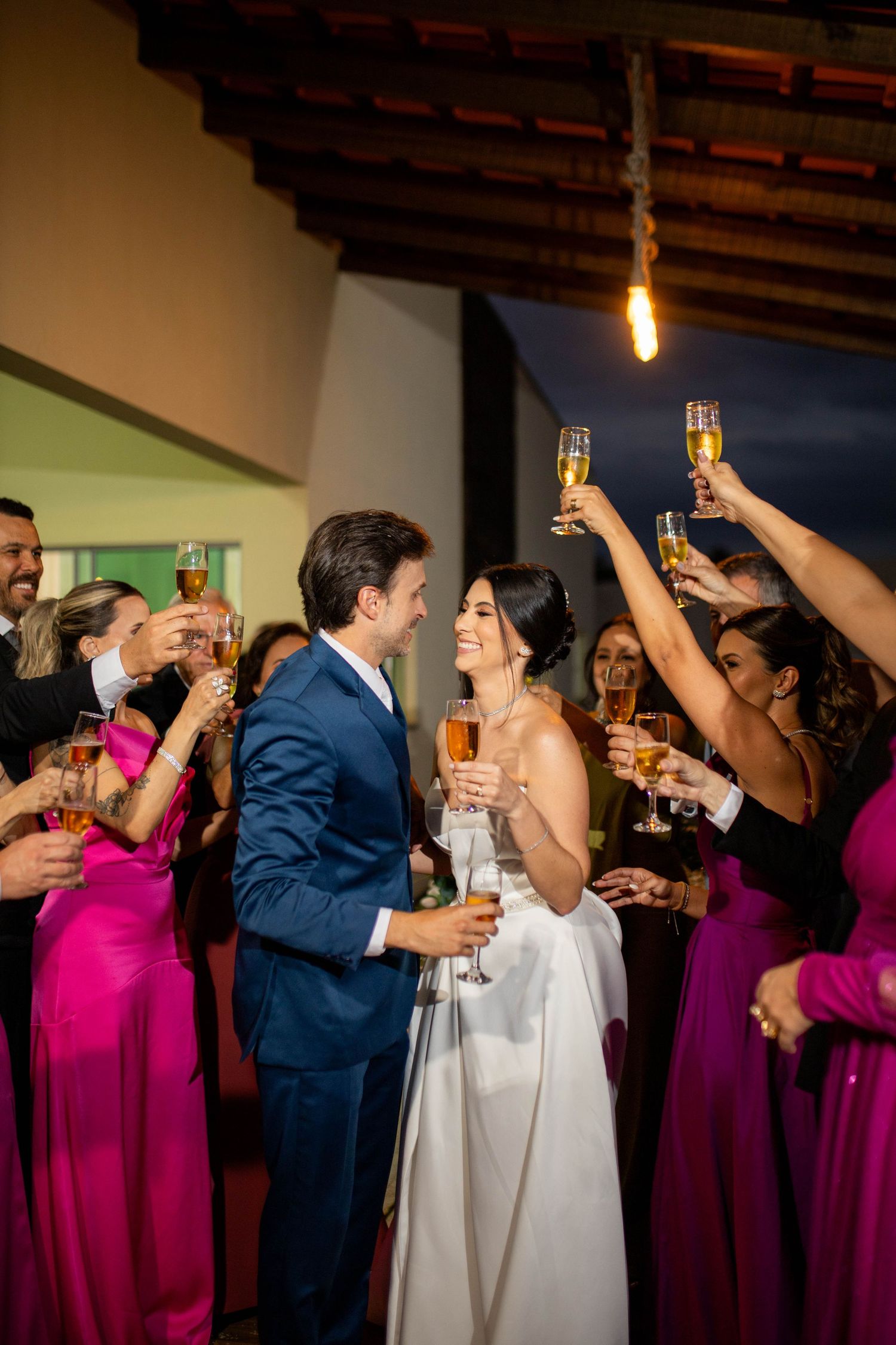 Bride and groom toast champagne with guests in pink gowns at elegant evening wedding reception celebration.