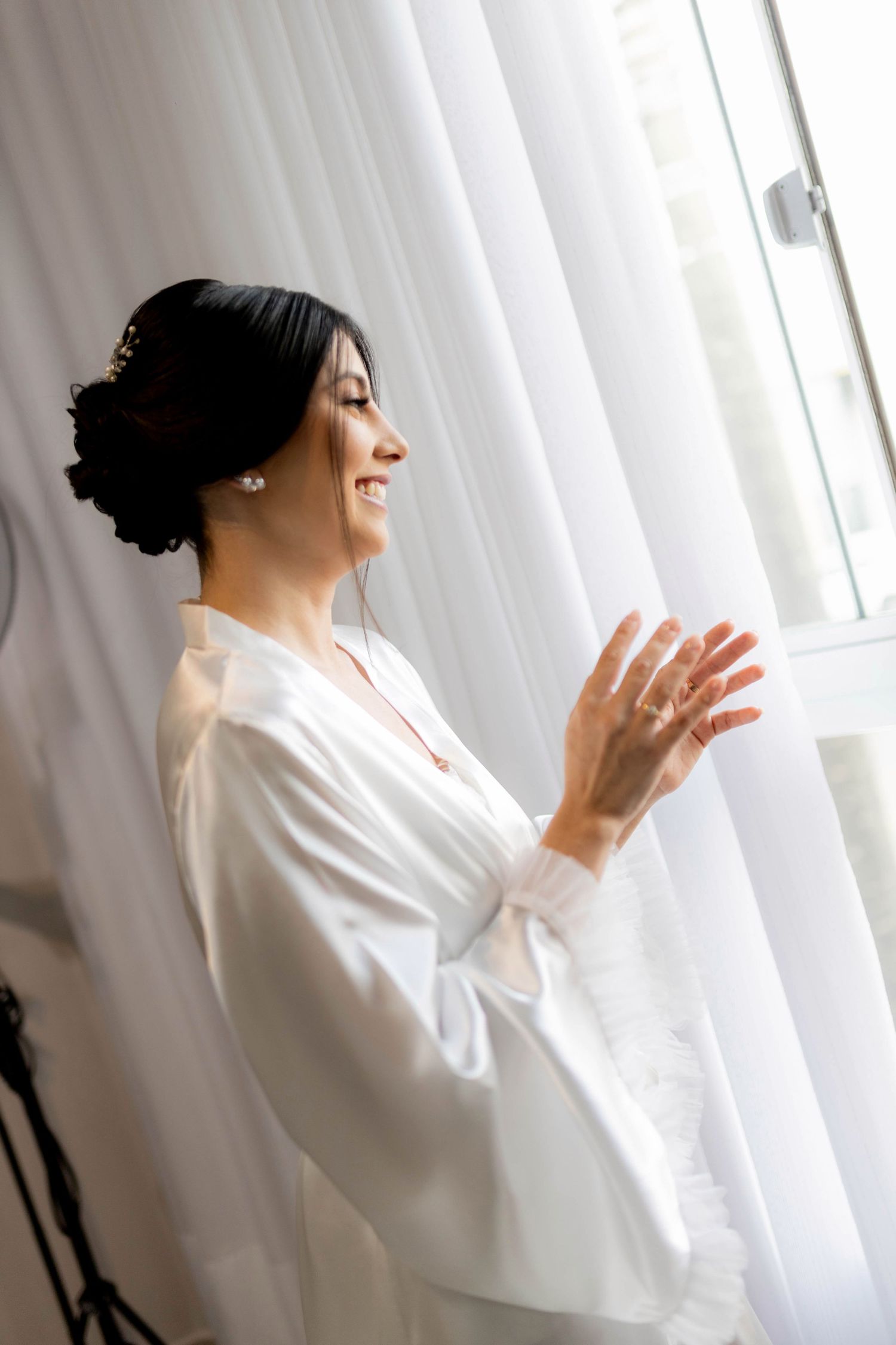Smiling bride in white robe standing by a sunlit window, wearing pearl earrings and elegant updo hairstyle.