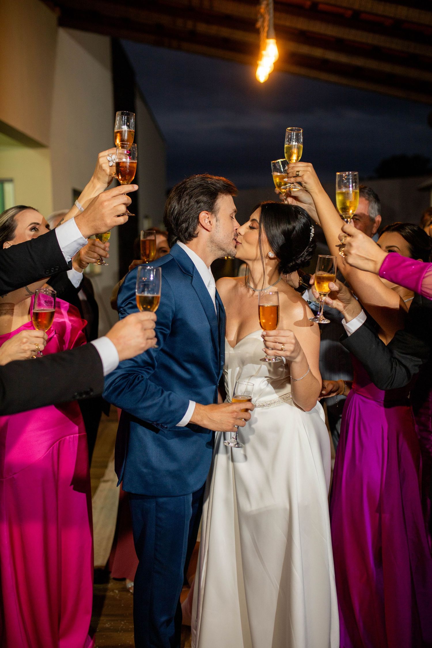 Newlyweds share a kiss while guests in pink gowns raise champagne glasses in celebration at an elegant evening wedding reception.