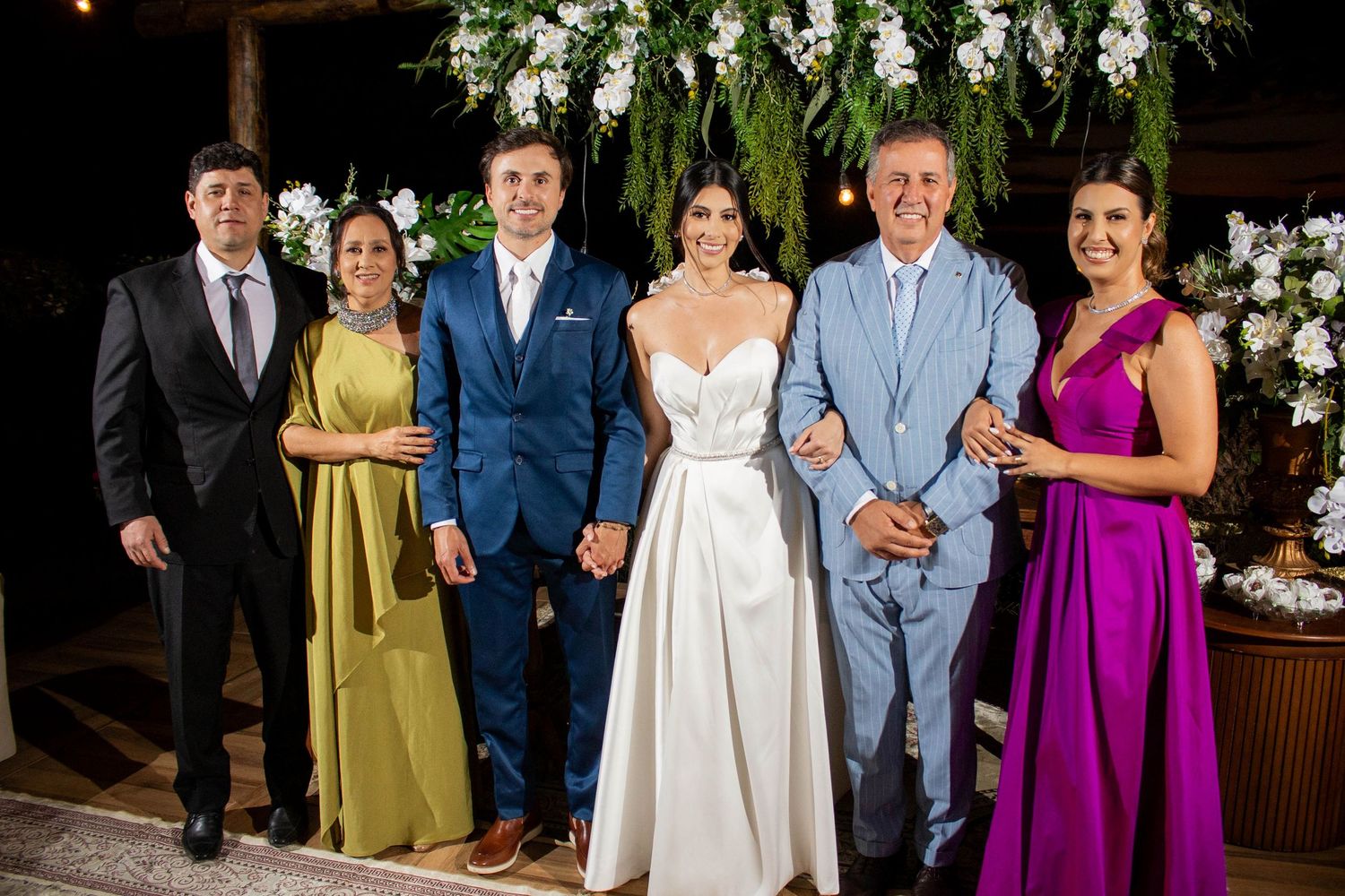 Wedding party of six posing together, bride in white gown, groom in blue suit, guests in formal attire outdoors.