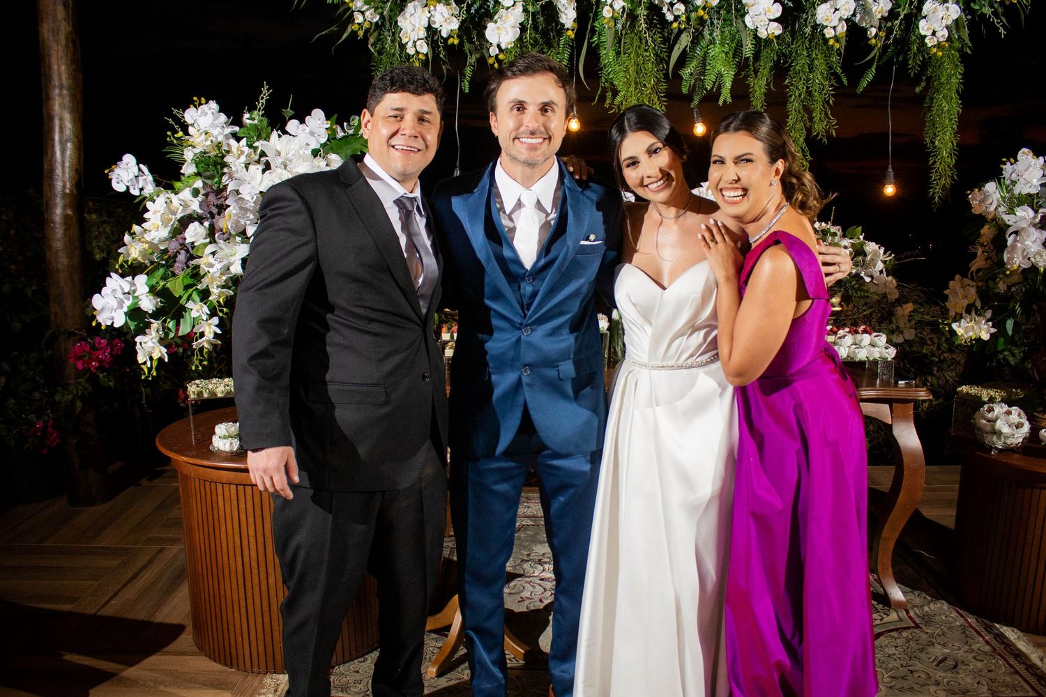 Four elegantly dressed guests pose together at a wedding reception, surrounded by lush floral arrangements and warm ambient lighting.