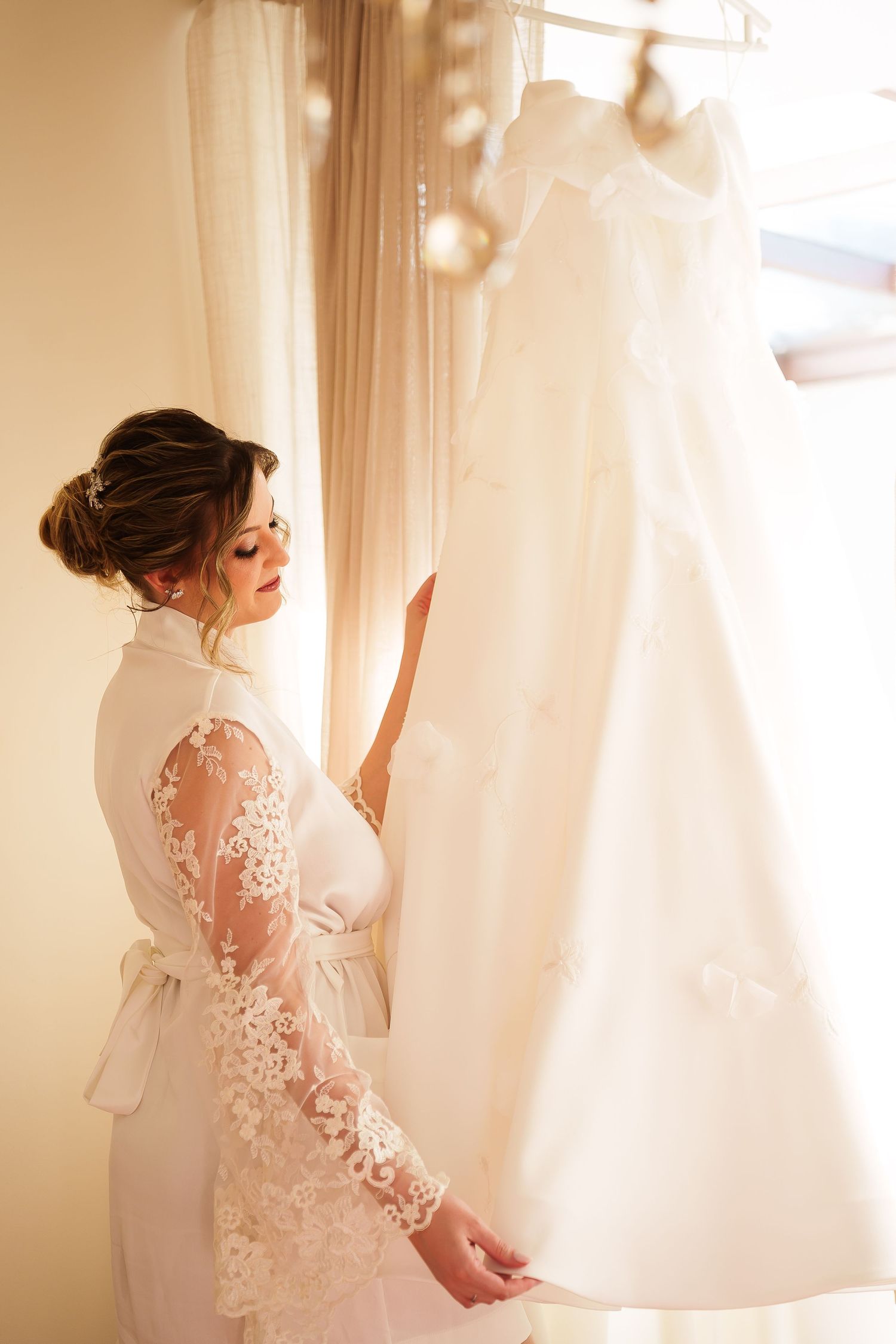 Bride in lace robe admiring her wedding dress hanging by a sunlit window on her wedding morning.