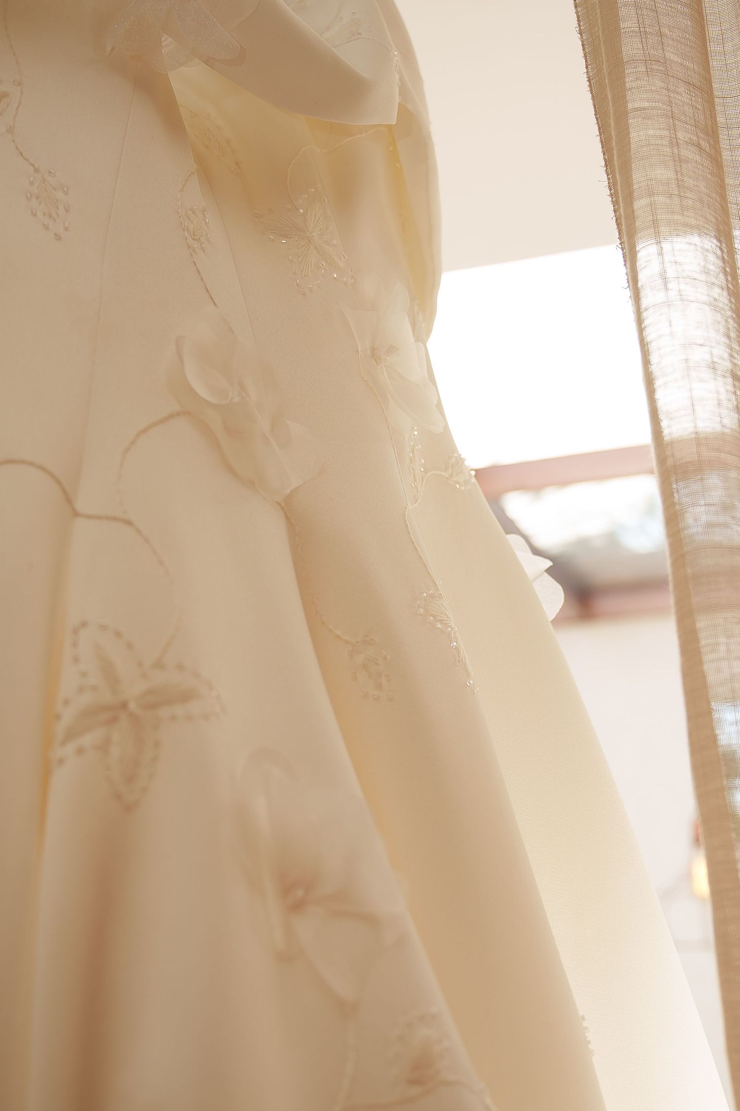Close-up of an elegant ivory wedding dress with floral embroidery, hanging near a sheer curtain in soft natural light.