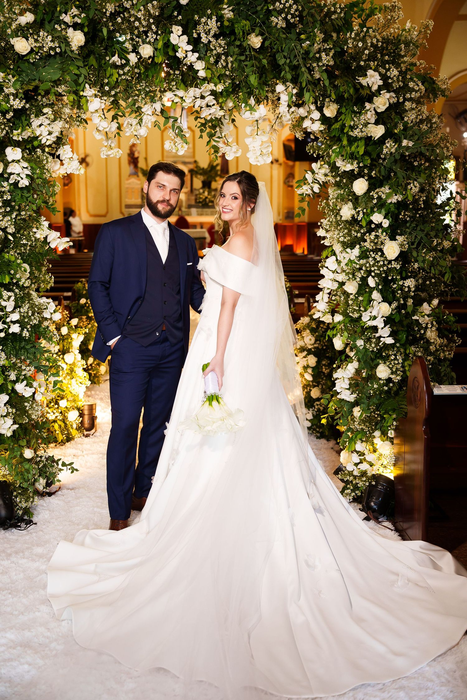 Bride and groom posing under a lush floral arch of white roses and greenery inside an elegant church.