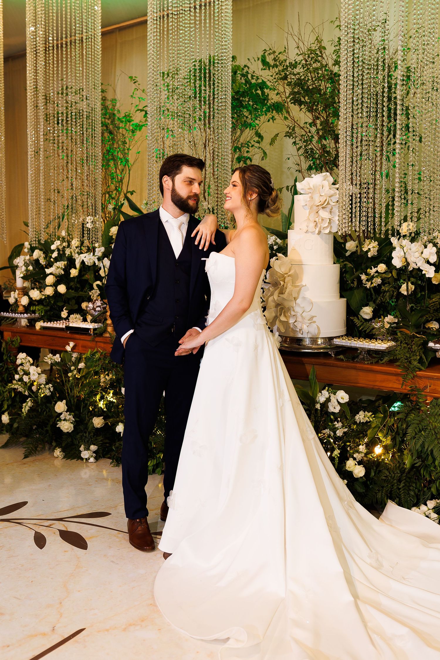 Elegant bride and groom pose together in front of a floral backdrop with a white wedding cake at their reception.