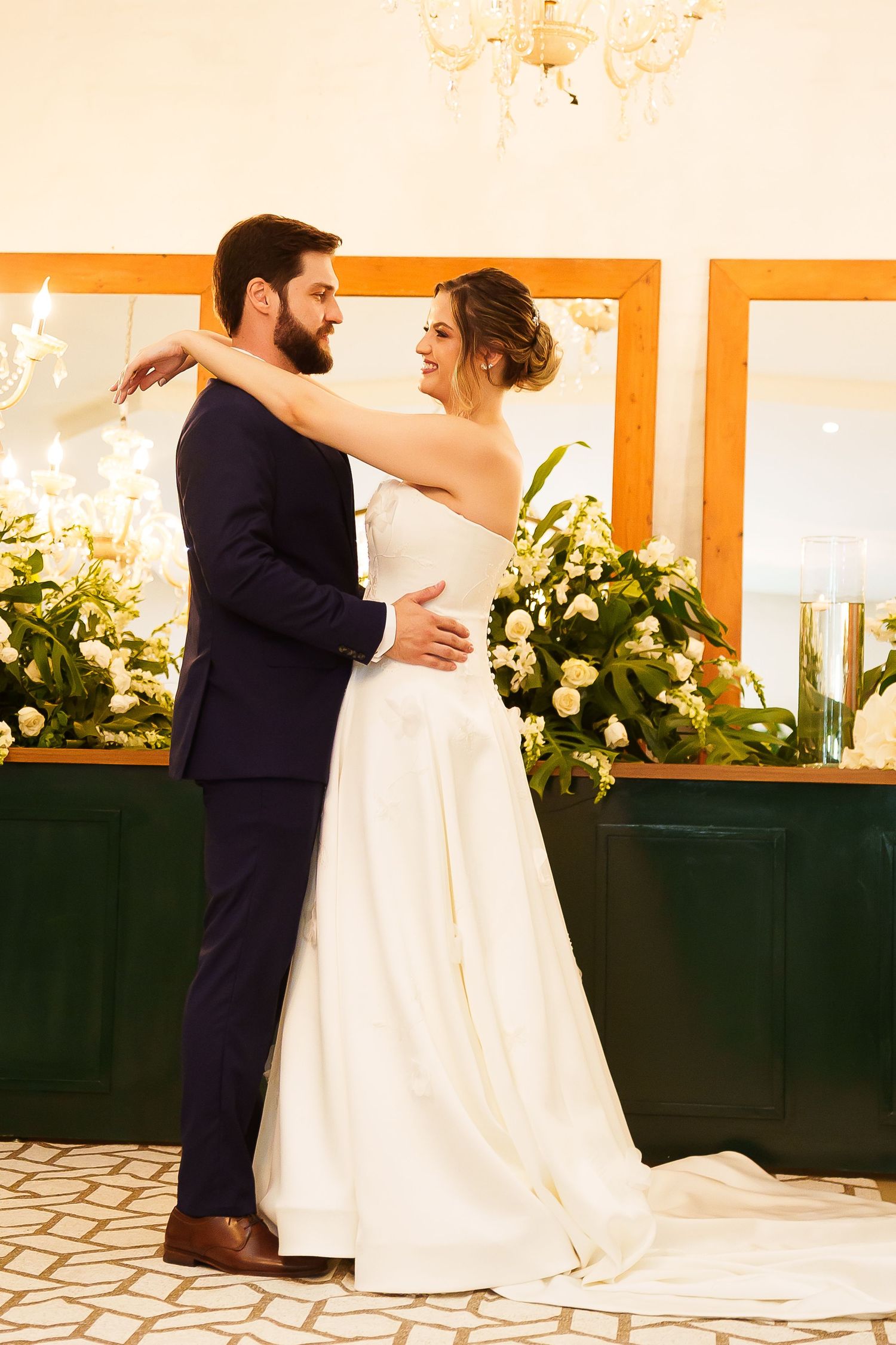 Wedding couple dancing elegantly, bride in white strapless gown with train, groom in navy suit, floral arrangement behind.