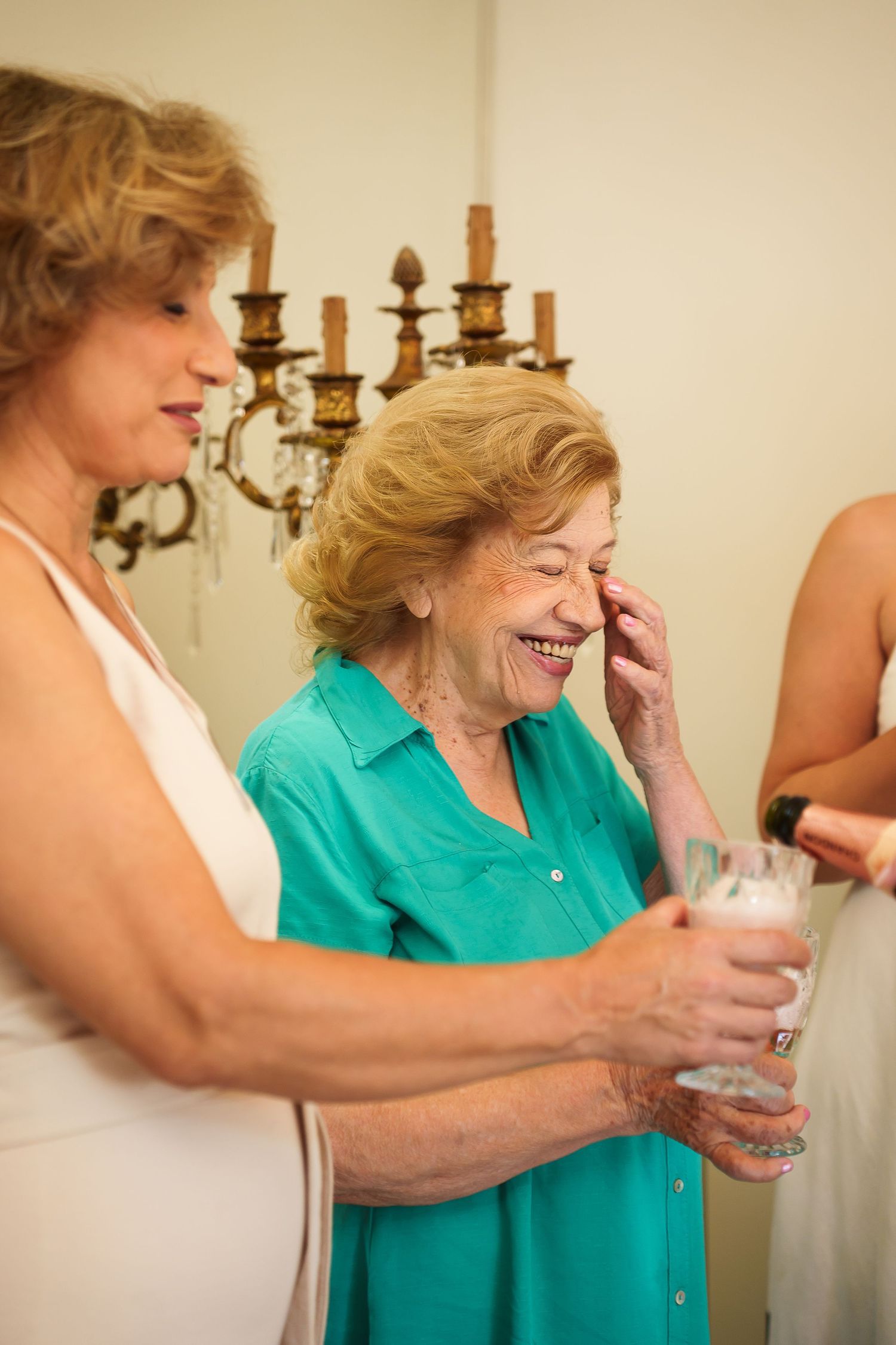 Elderly woman in teal shirt laughing joyfully while holding a glass at a celebration with friends.