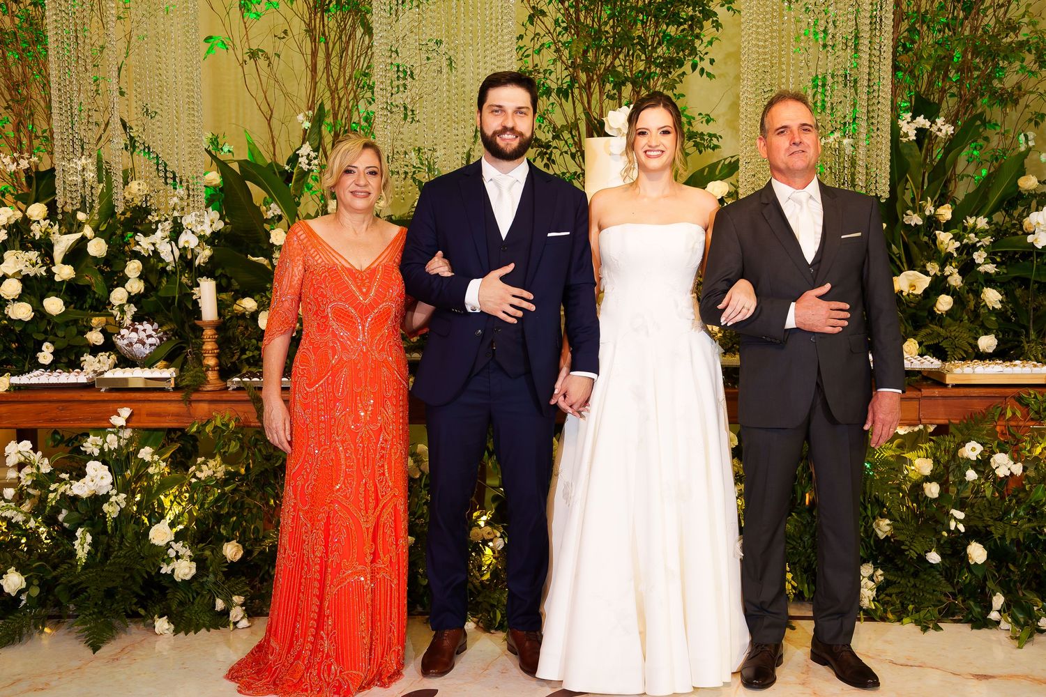 Bride and groom pose with family members at elegant wedding reception with floral greenery backdrop.