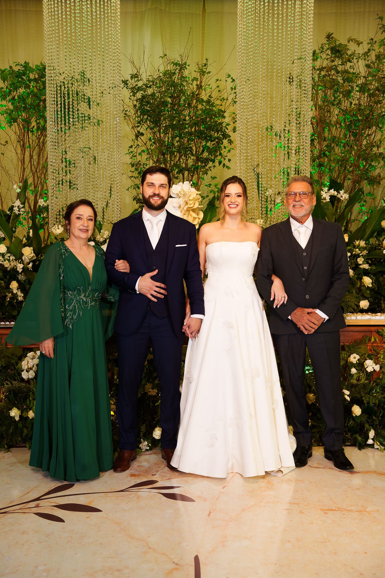 Bride and groom pose with family members at elegant wedding reception with floral backdrop and greenery.