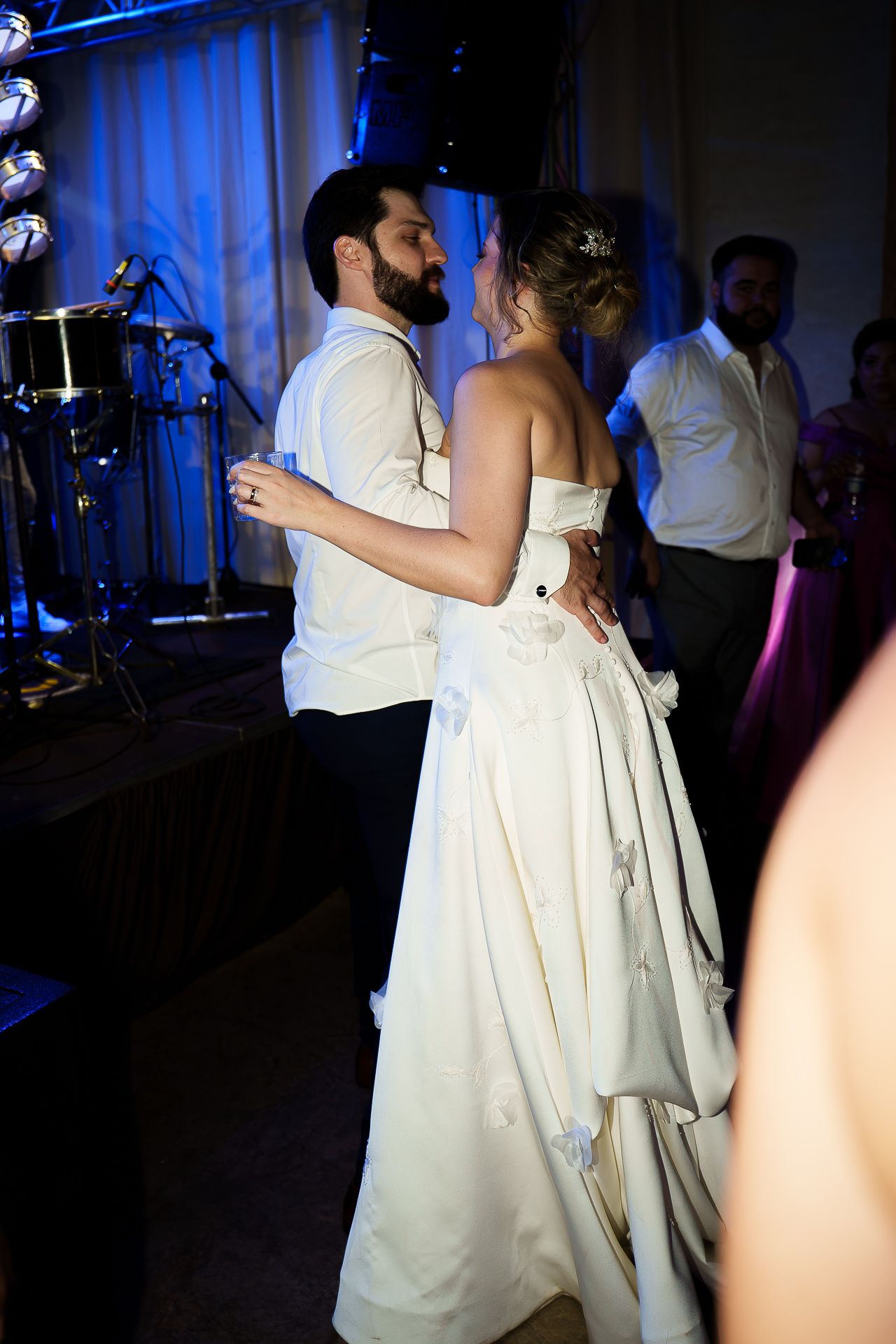 Bride and groom share their first dance at wedding reception, surrounded by guests under blue ambient lighting.