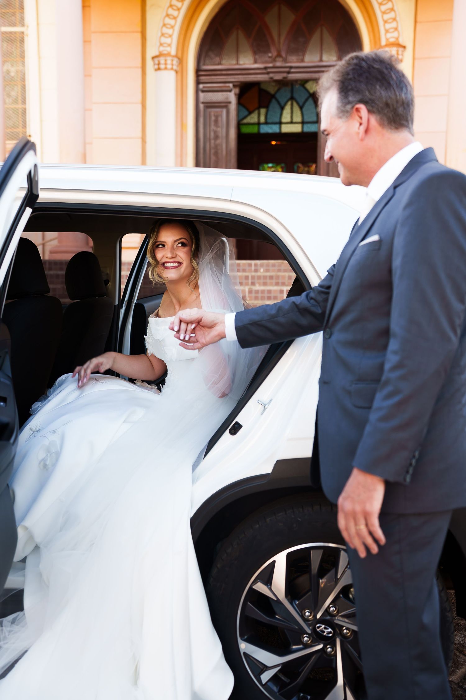 Smiling bride in white gown exits white limo, assisted by man in dark suit, outside a church.